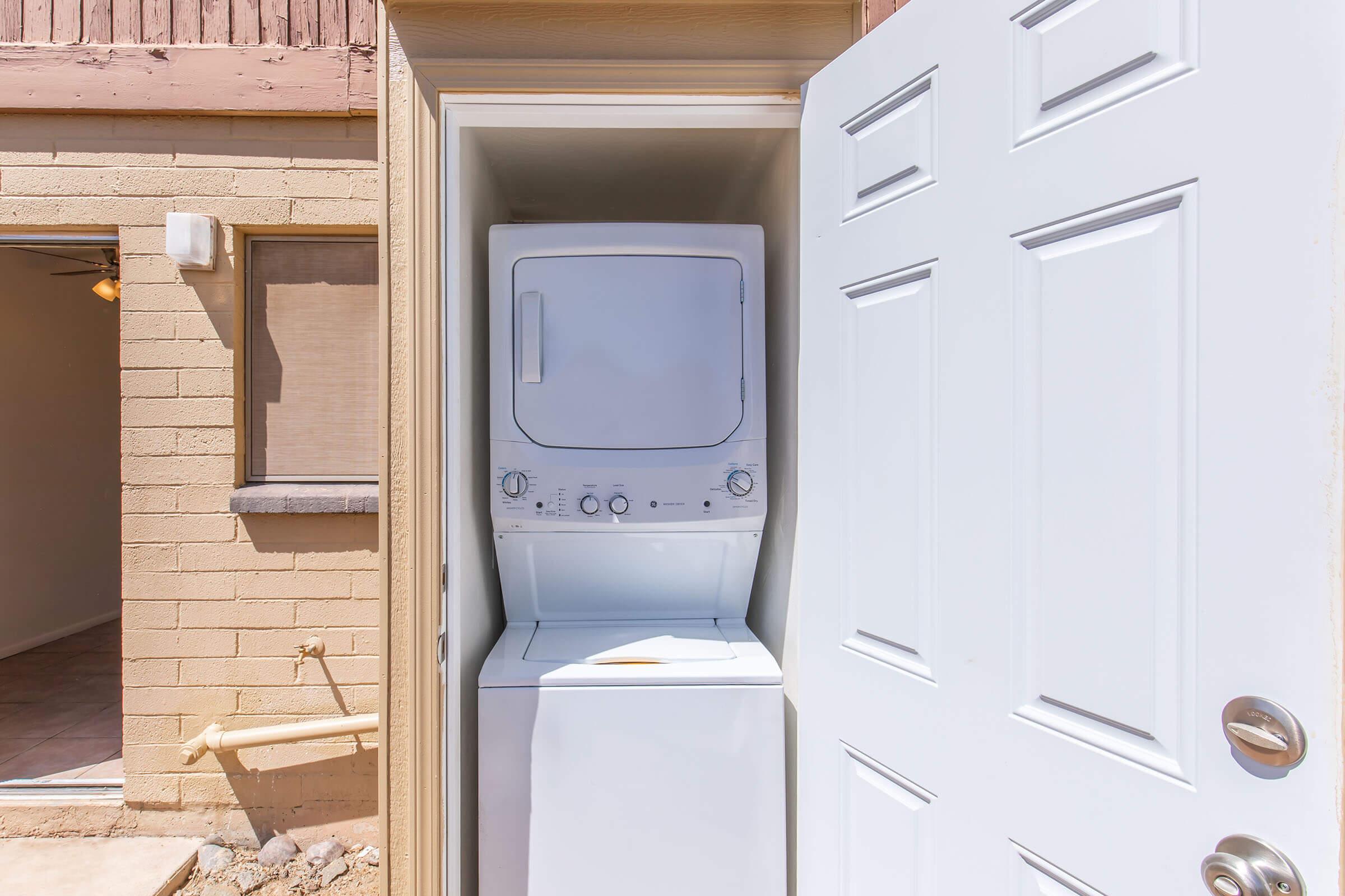 A compact laundry area featuring a stacked washer and dryer unit inside a small closet. The closet door is open, revealing the appliances. The surrounding walls are beige, and there are light fixtures visible in the background. Sunlight enters the area, providing a bright and inviting atmosphere.