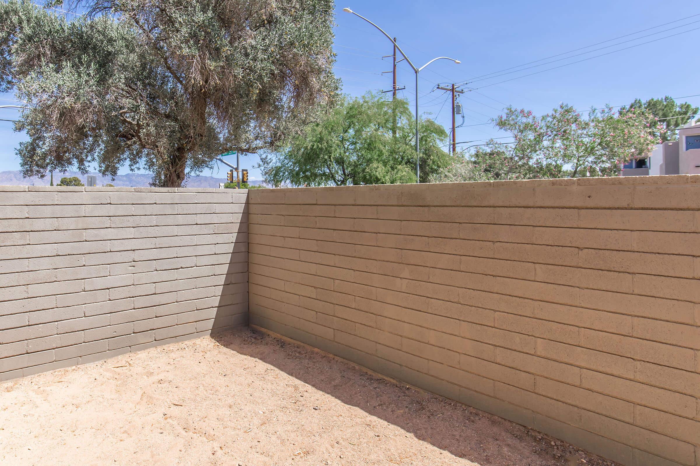 A corner view of an outdoor area featuring two gray concrete walls, with a dry sandy ground. In the background, there are trees and blue sky, along with some power lines visible. The setting appears to be a tranquil and empty space, likely intended for a garden or a patio.