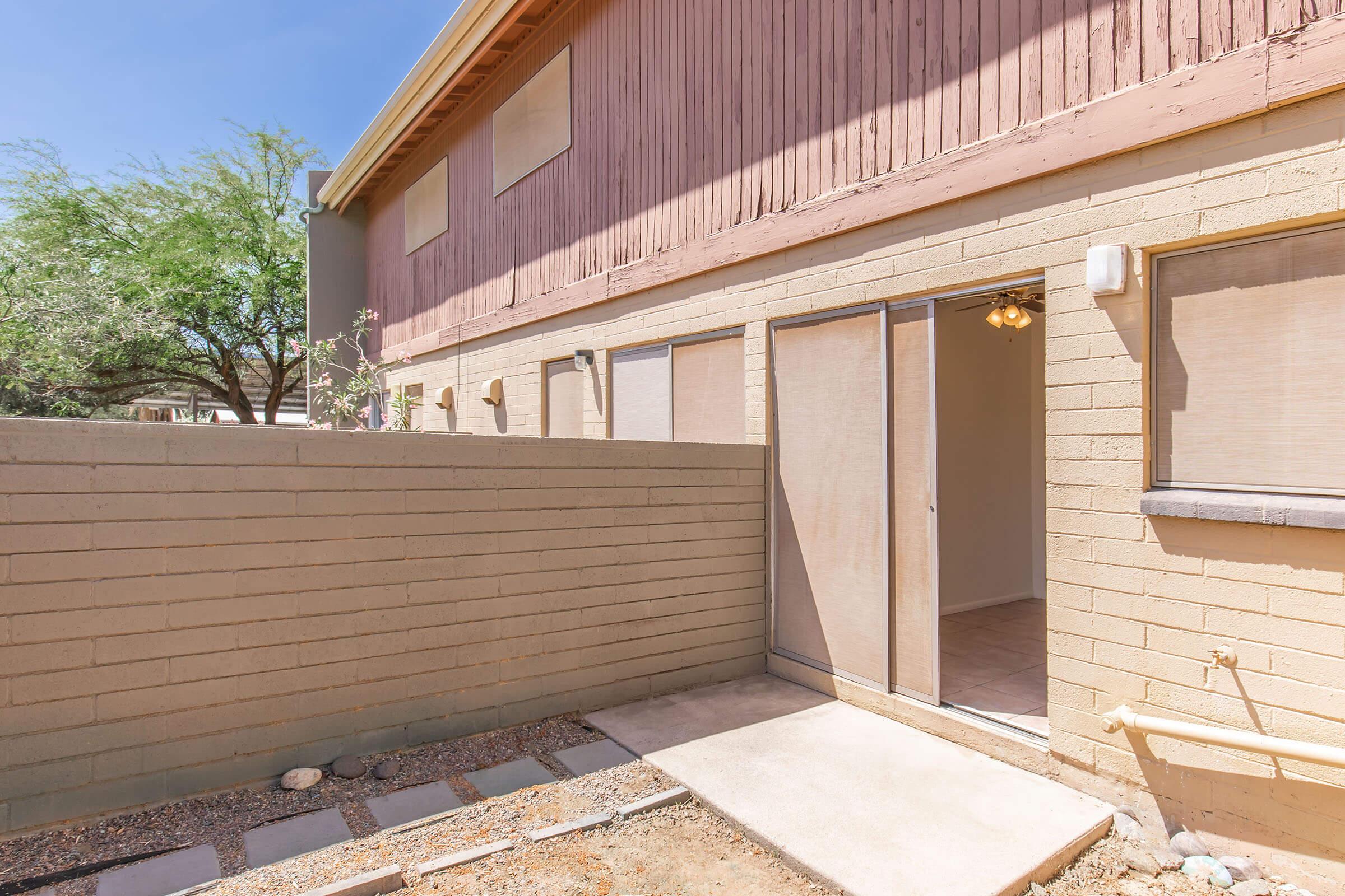 Exterior view of a ground-level entrance to a building with a light-colored brick wall and a small pathway. The door is partially open, revealing an empty interior space. There are windows on the side, and the surrounding area features desert-like landscaping with minimal vegetation. Clear blue sky overhead.