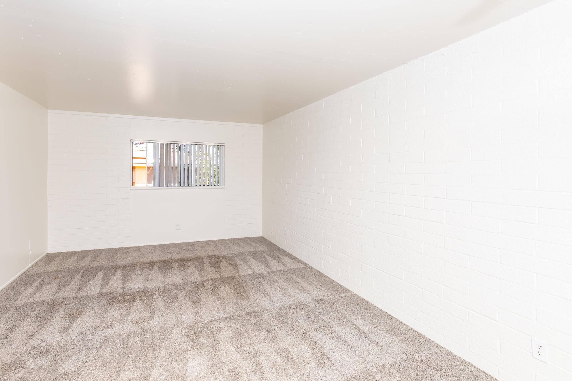 A spacious, empty room with white brick walls and a light-colored carpet. A small window is visible on one wall, allowing natural light to enter. The room has no furniture, creating a minimalist and open feel.