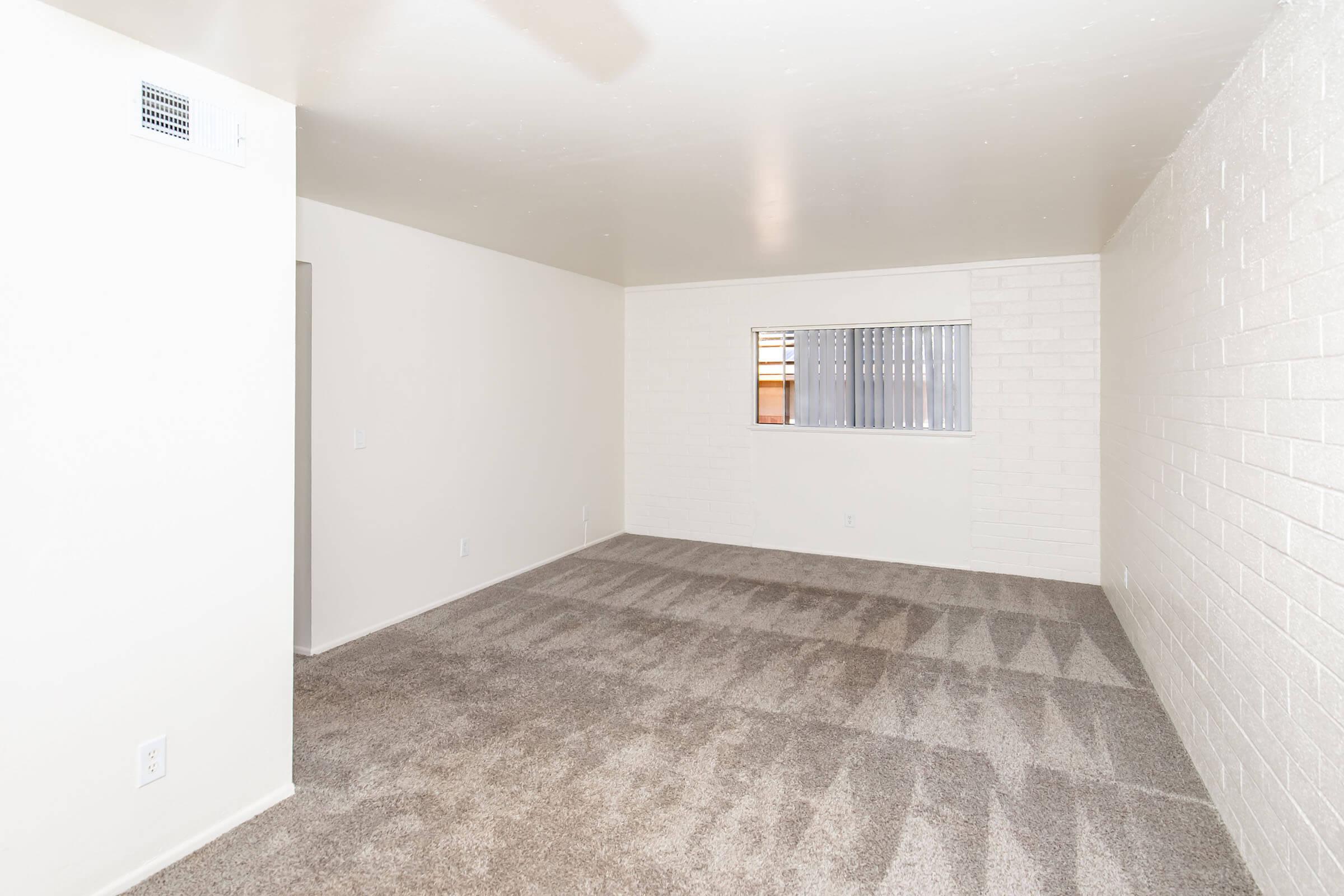 A spacious, empty room with light-colored walls and plush carpet. A window with blinds allows natural light to enter. The room features clean lines and minimal decor, making it suitable for various uses like a living area or bedroom.