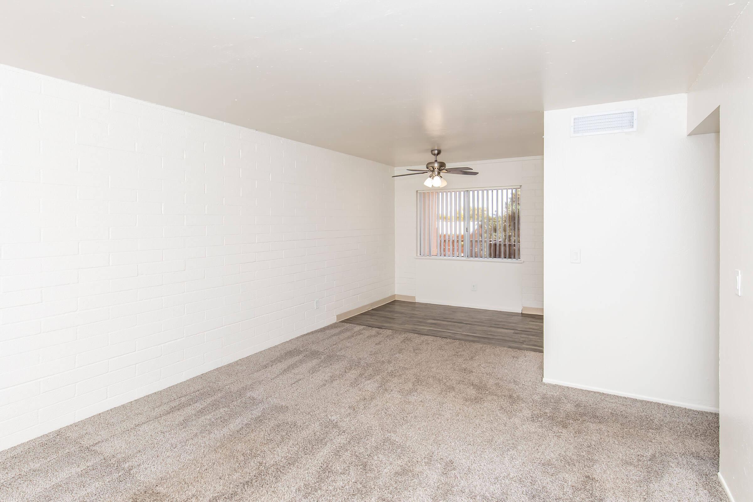 An empty living room with light-colored walls, a ceiling fan, and a large window with blinds. The floor is covered in beige carpeting, while the adjacent area features laminate flooring. The space has a bright and open feel, with natural light coming in through the window.