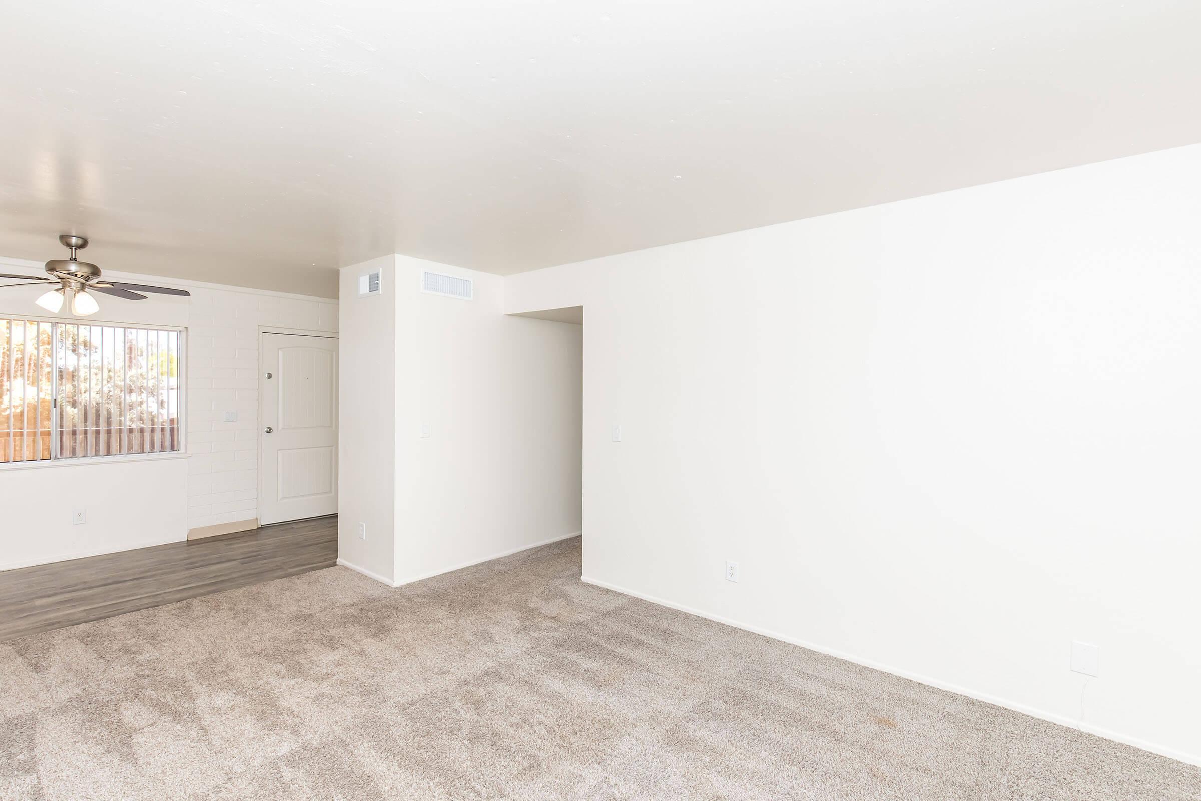 A spacious, empty living area featuring light-colored walls and carpet. A ceiling fan is visible, and there's a window providing natural light. An entrance door is located on the left, with a small hallway leading to another room. The overall ambiance is bright and airy, with a minimalist design.