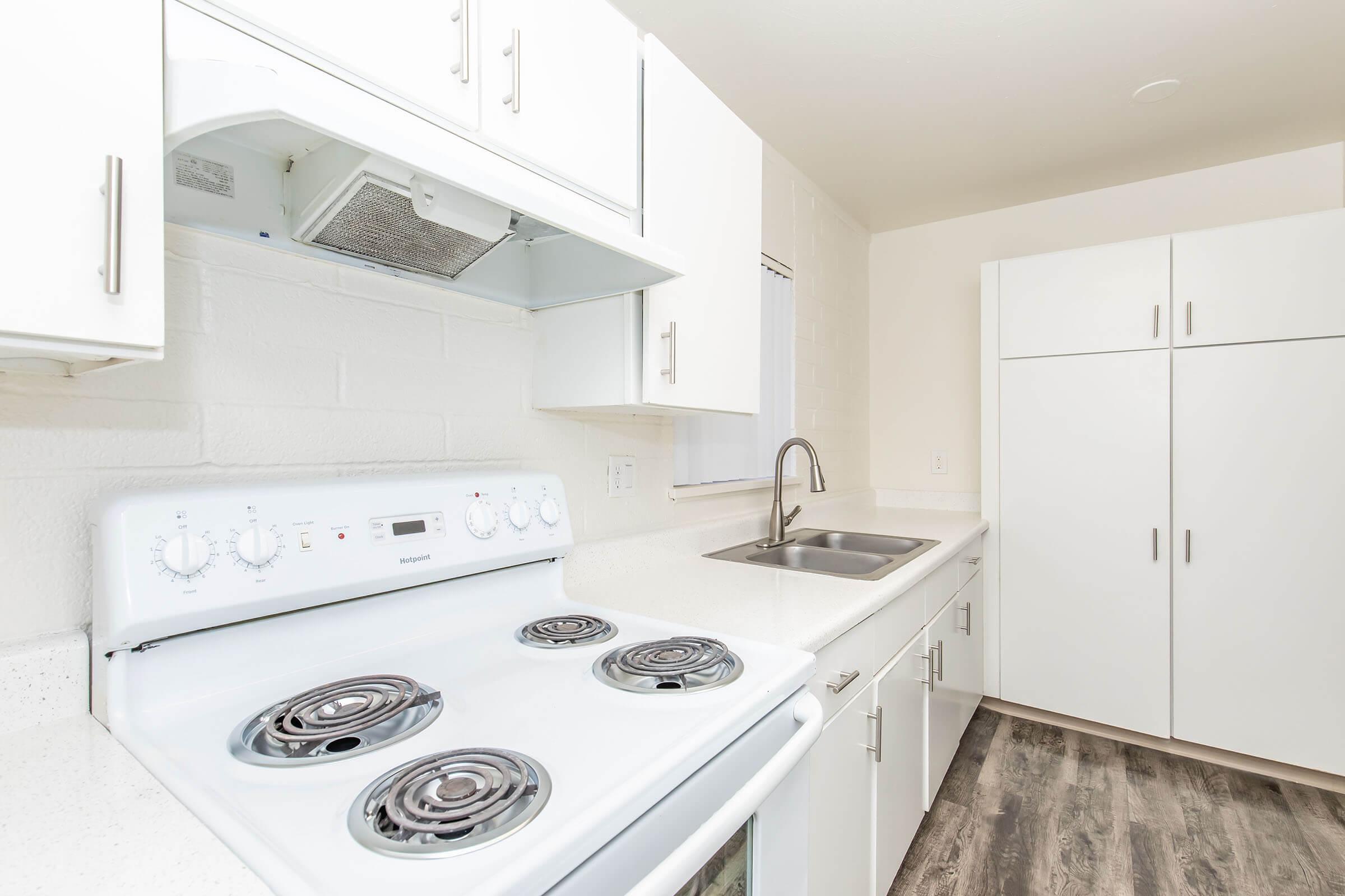 A clean, modern kitchen featuring white cabinets, a white stove with burners, a stainless steel sink, and a refrigerator. The walls are light-colored, and the flooring is a dark, wood-like laminate. Natural light is coming through a window, creating a bright and airy atmosphere.
