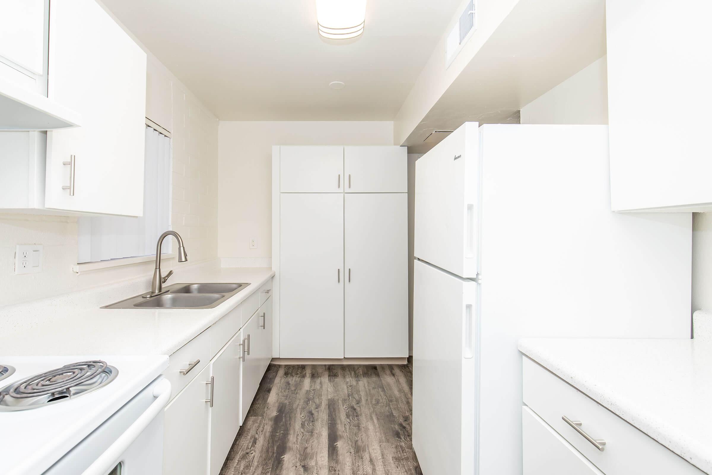 Modern white kitchen featuring sleek cabinets, a double sink, and a large refrigerator. The room has a minimalist design with light-colored countertops and wooden flooring. A window allows natural light to brighten the space, creating an inviting atmosphere.