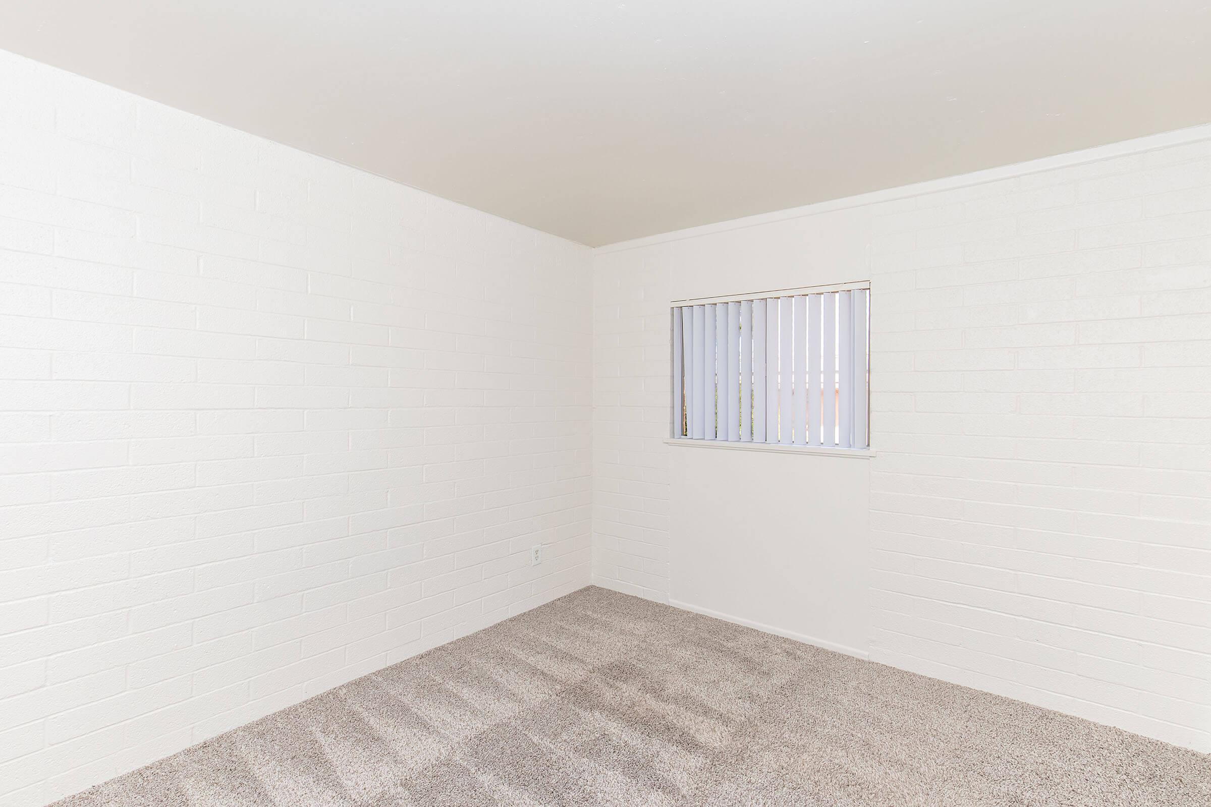 An empty room with white brick walls and a small window with vertical blinds. The floor is covered with light-colored carpet, and the overall space is well-lit, giving a clean and minimalist appearance. The room is devoid of furniture or decorations.