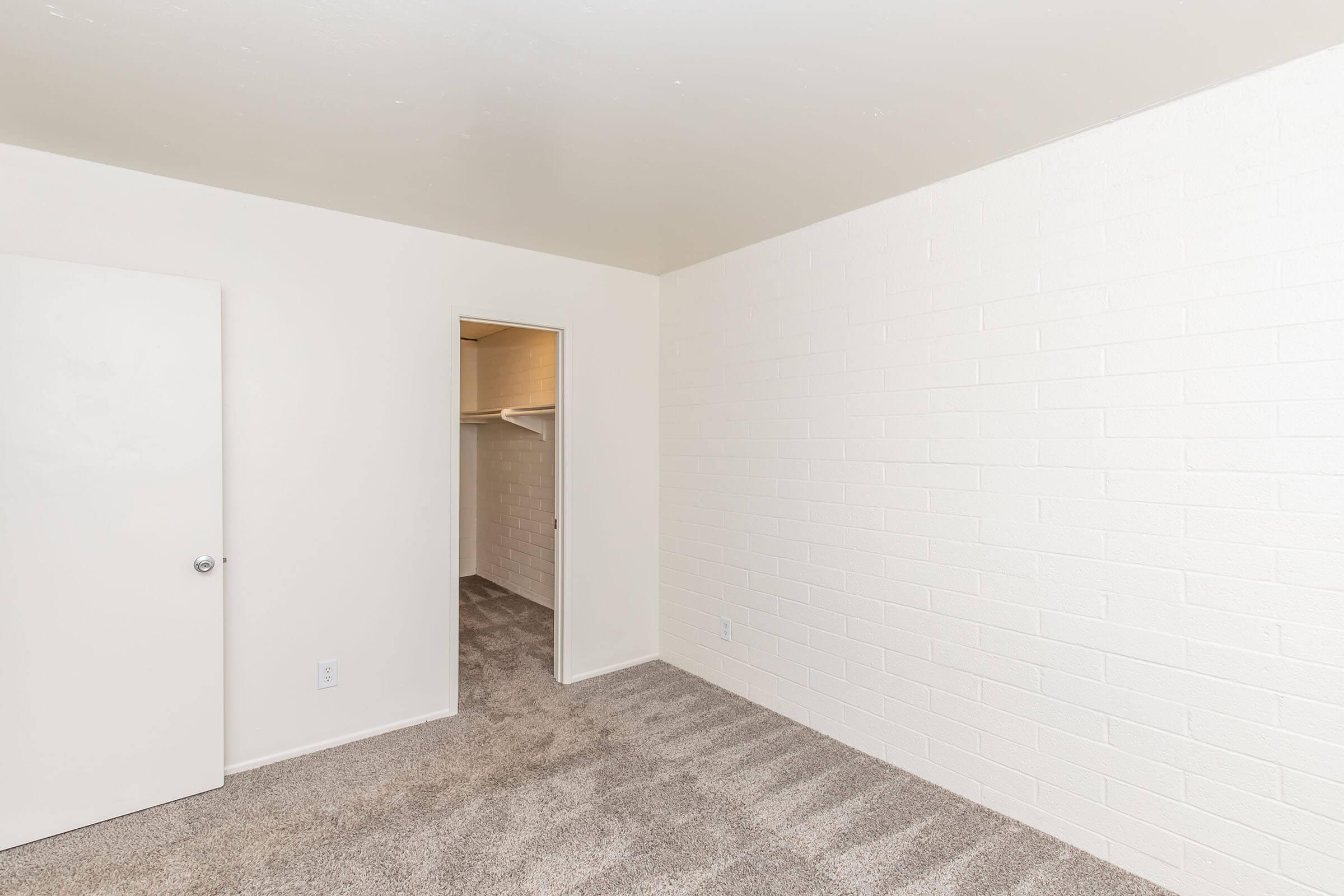 A vacant room with light-colored walls and beige carpet. There is a closed white door on the left and an open door leading to a closet on the right. The room is well-lit and spacious, with no furniture or decorations.