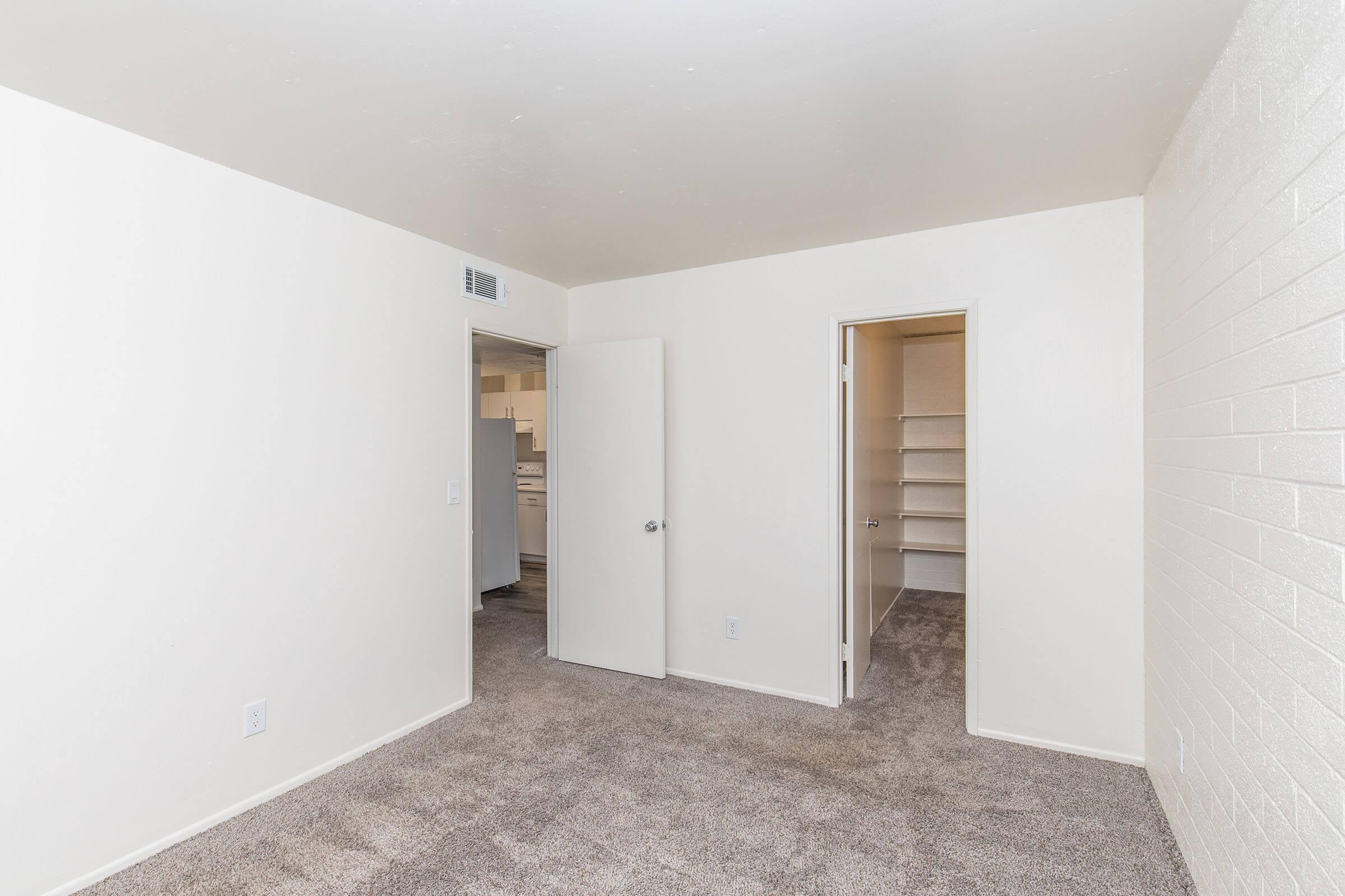A spacious, empty room with beige carpet and white walls. There are two doors: one leads to a closet with shelves, and the other appears to lead to a kitchen area. Natural light illuminates the room, creating a bright and inviting atmosphere.