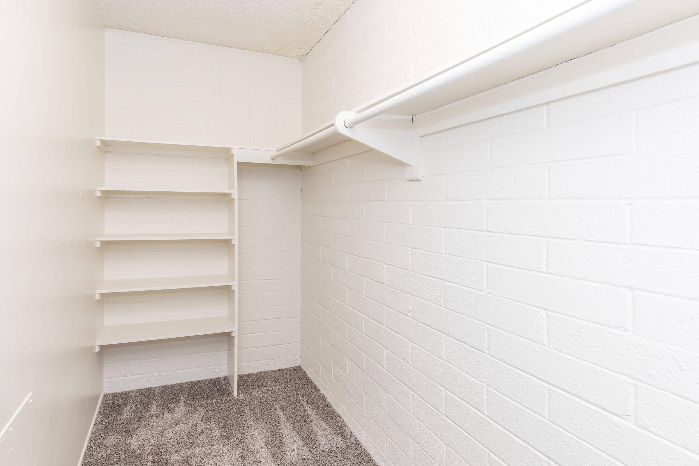 An empty closet with white brick walls, featuring a hanging rod and four white shelves along one side. The floor is covered in a soft, gray carpet.