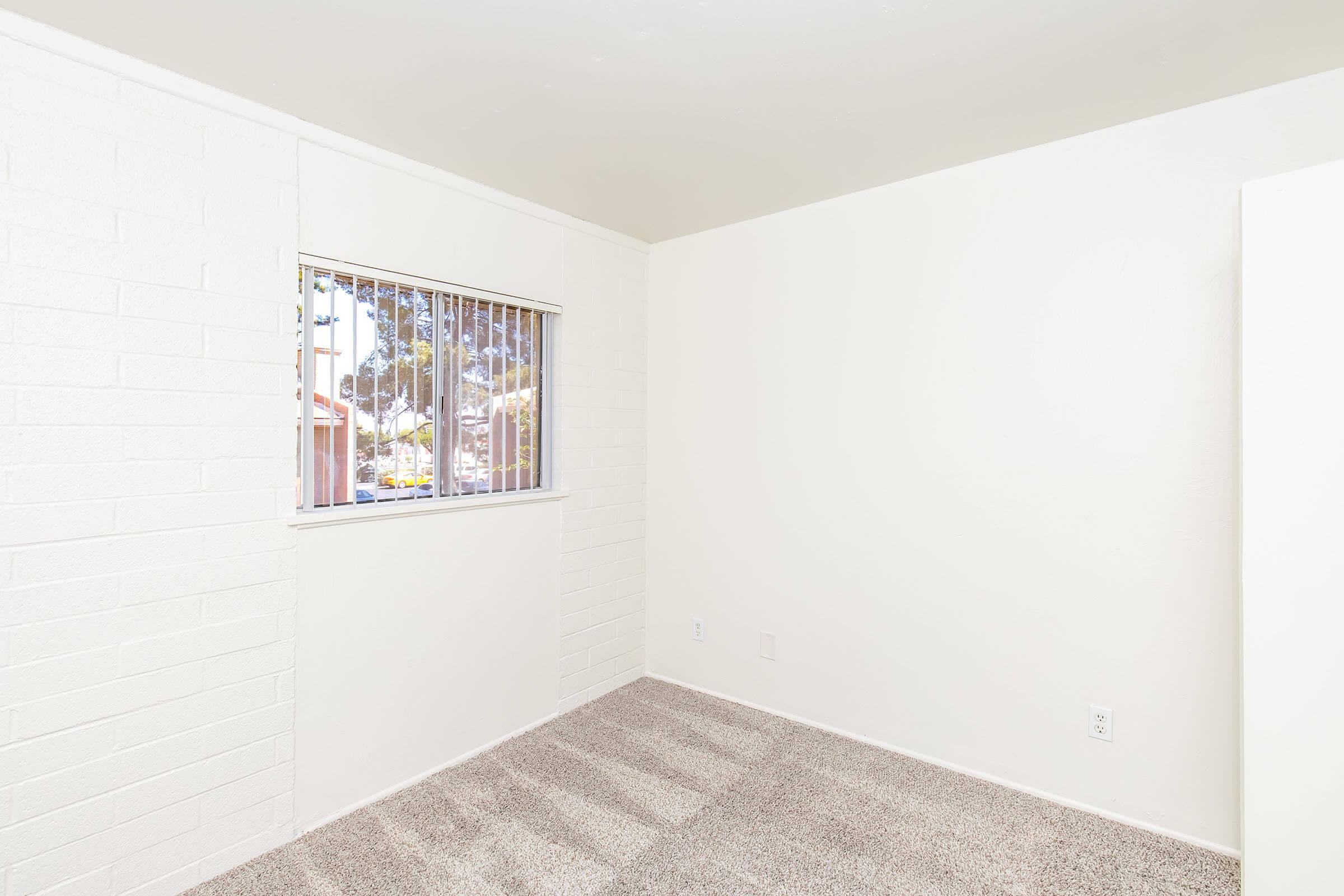 Empty room with white brick walls and a window featuring vertical blinds. The floor is covered in light gray carpet, and there is minimal furniture. Natural light enters the space through the window, illuminating the area.