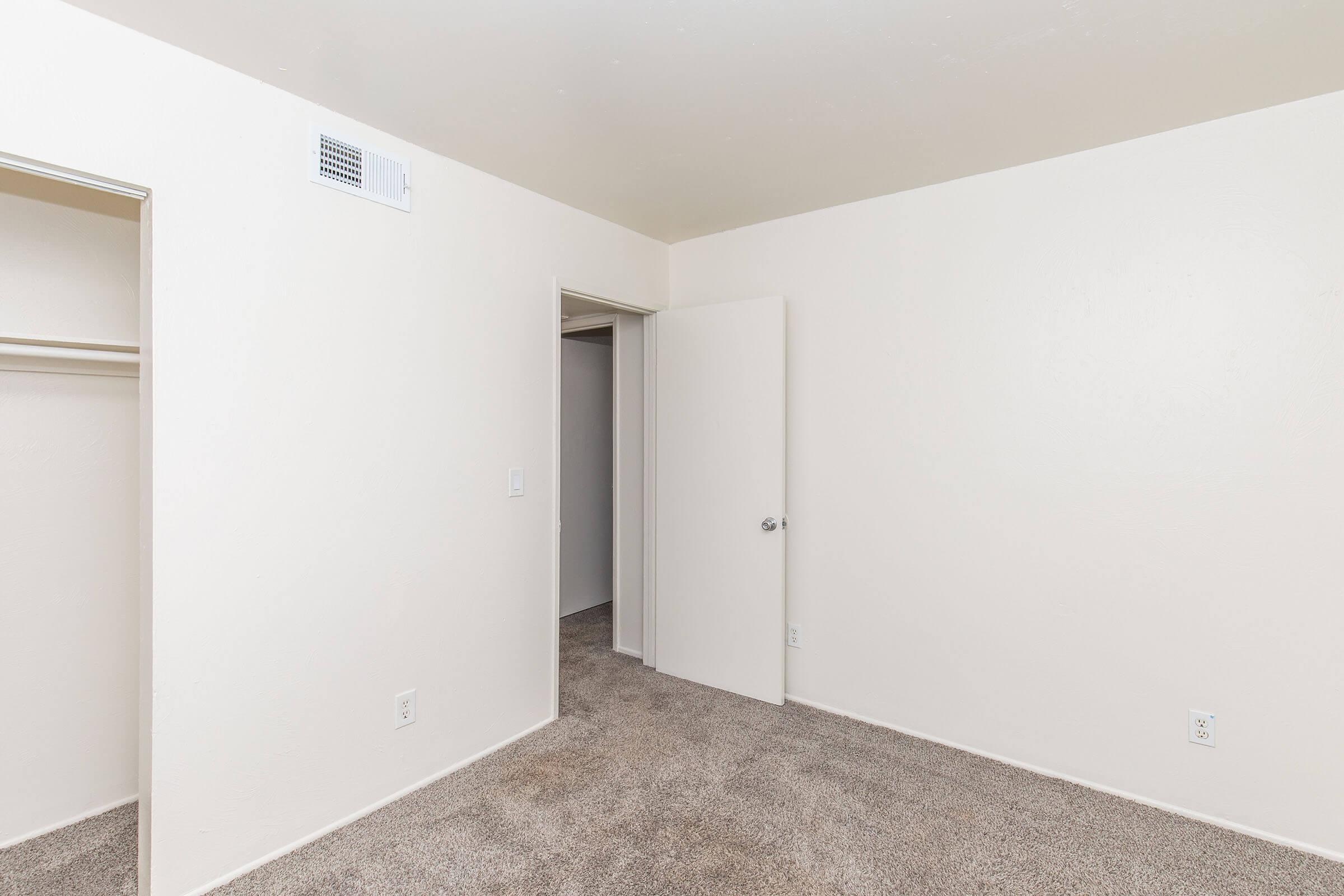 A vacant room with light beige walls and gray carpet. There is a doorway leading to another room, and an open closet on the left side. The room is well-lit and has a minimalistic appearance, with no furniture or decorations.