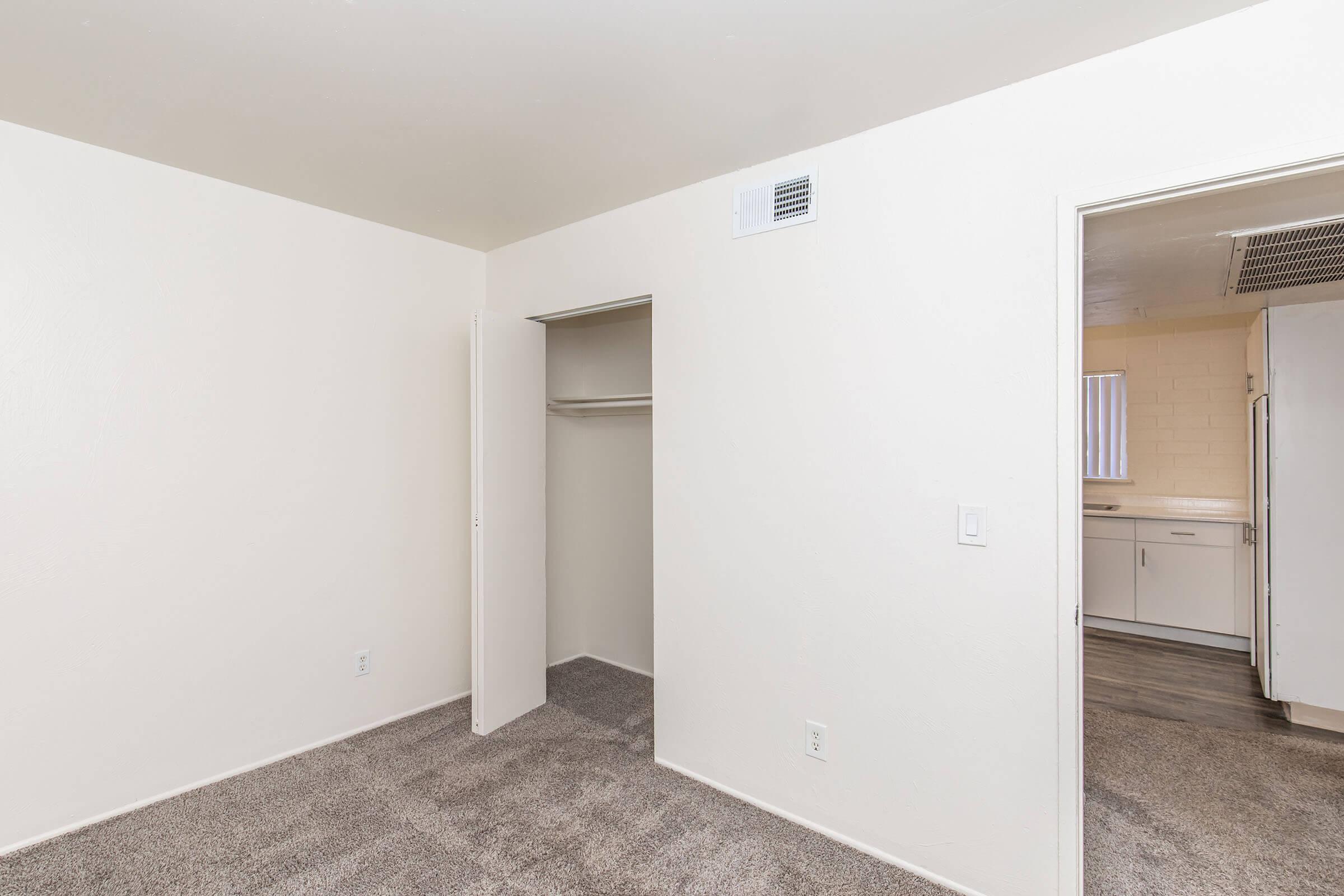 A well-lit, empty room with beige walls and carpeted flooring. On one side, there is an open closet with a sliding door. A doorway leads to another room visible in the background, suggesting this is part of a larger living space. The overall feel is clean and spacious.