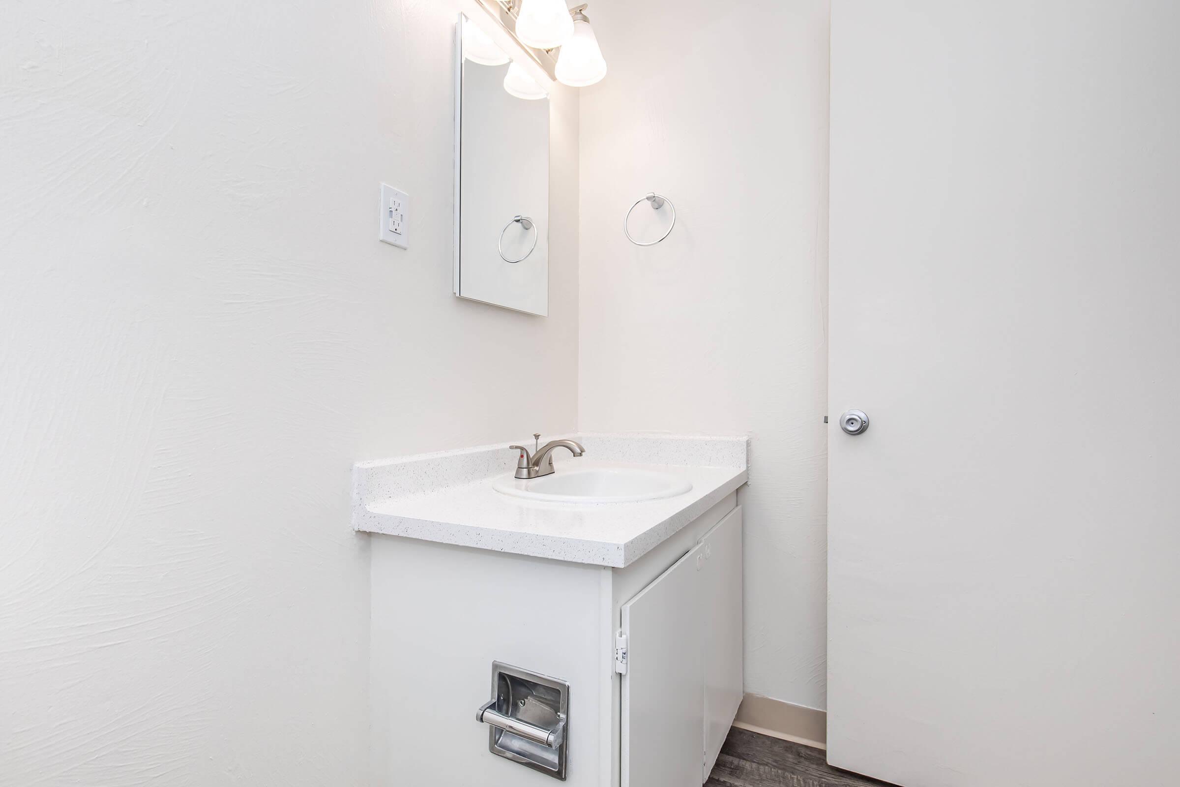 A simple bathroom setting featuring a white countertop sink with a modern faucet, a single mirror mounted above it, a towel holder on the wall, and a closed door on the right. The walls are painted in a light color, creating a bright and clean atmosphere.