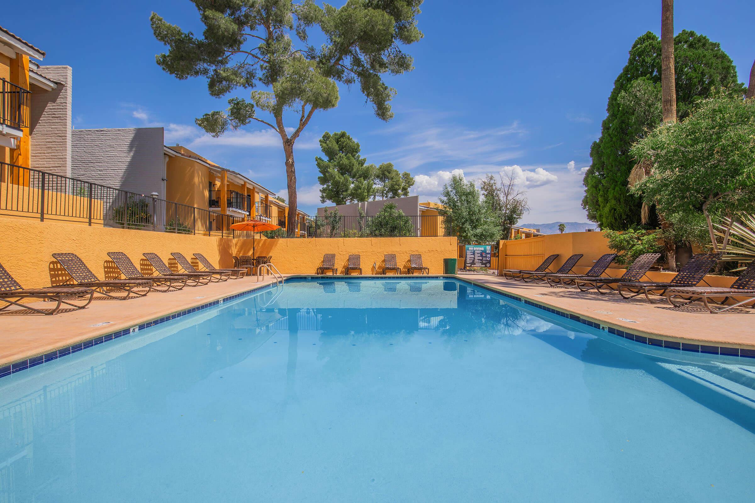A clear swimming pool surrounded by lounge chairs under a bright blue sky. In the background, there are yellow buildings and trees, creating a relaxing outdoor atmosphere.