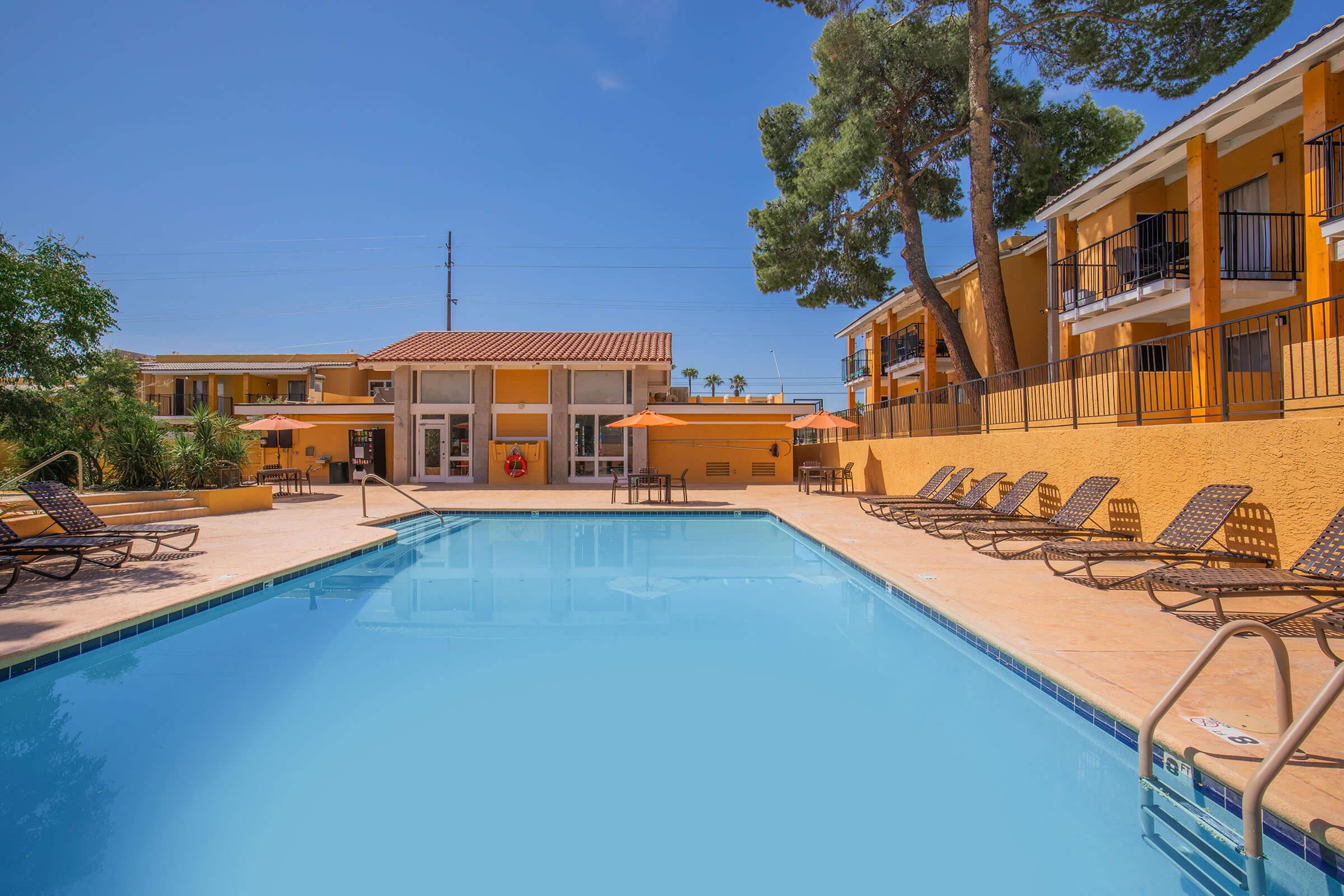 A bright, sunny view of a pool area at a hotel or resort, featuring a clear blue swimming pool surrounded by lounge chairs. The backdrop includes multiple buildings painted in warm colors, with trees providing some shade. Umbrellas are set up near the poolside.