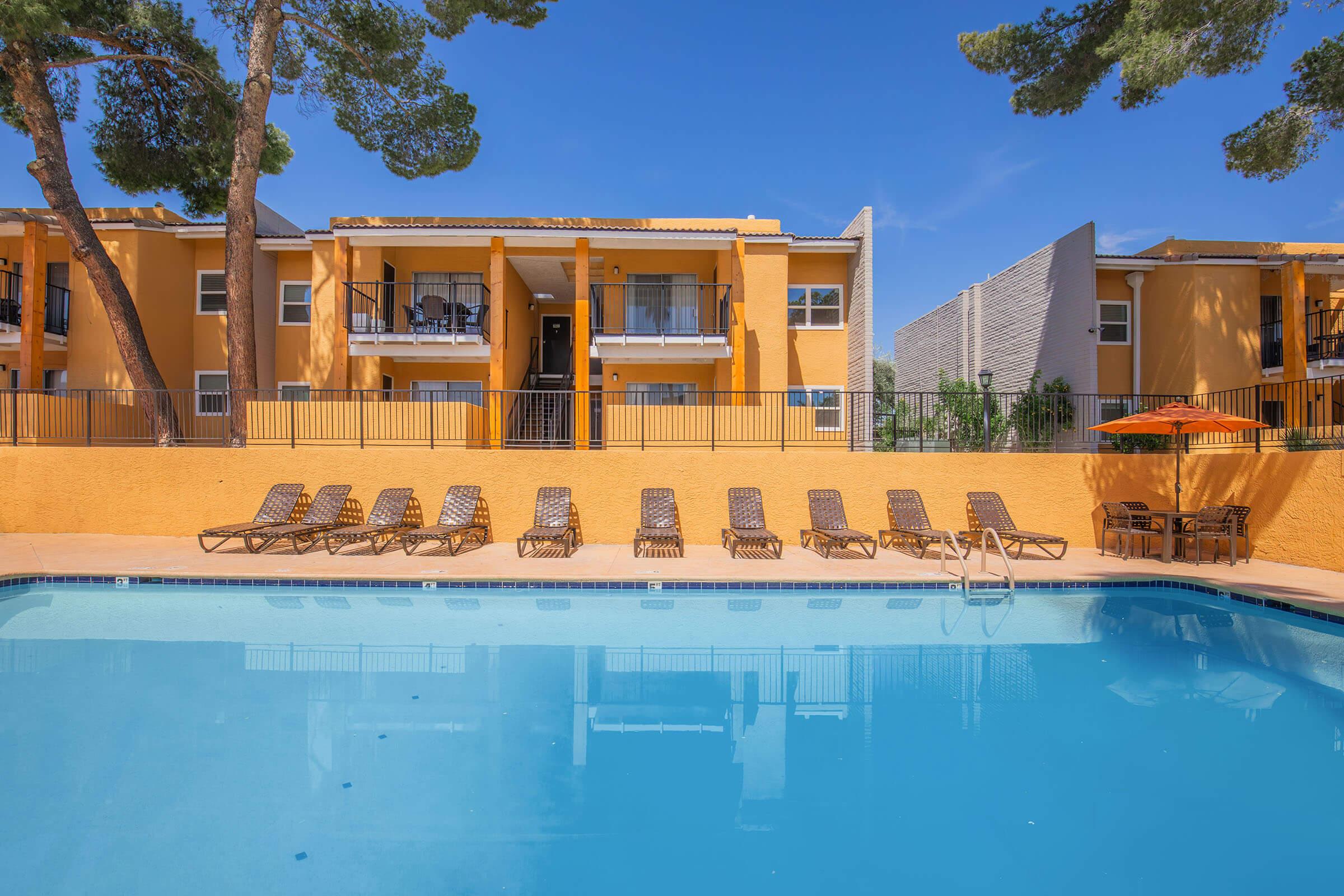 A sparkling blue swimming pool surrounded by loungers, with orange apartment buildings in the background. Tall pine trees provide shade, and an orange umbrella offers additional seating near the pool area, creating a relaxing outdoor environment. Bright blue sky complements the scene.