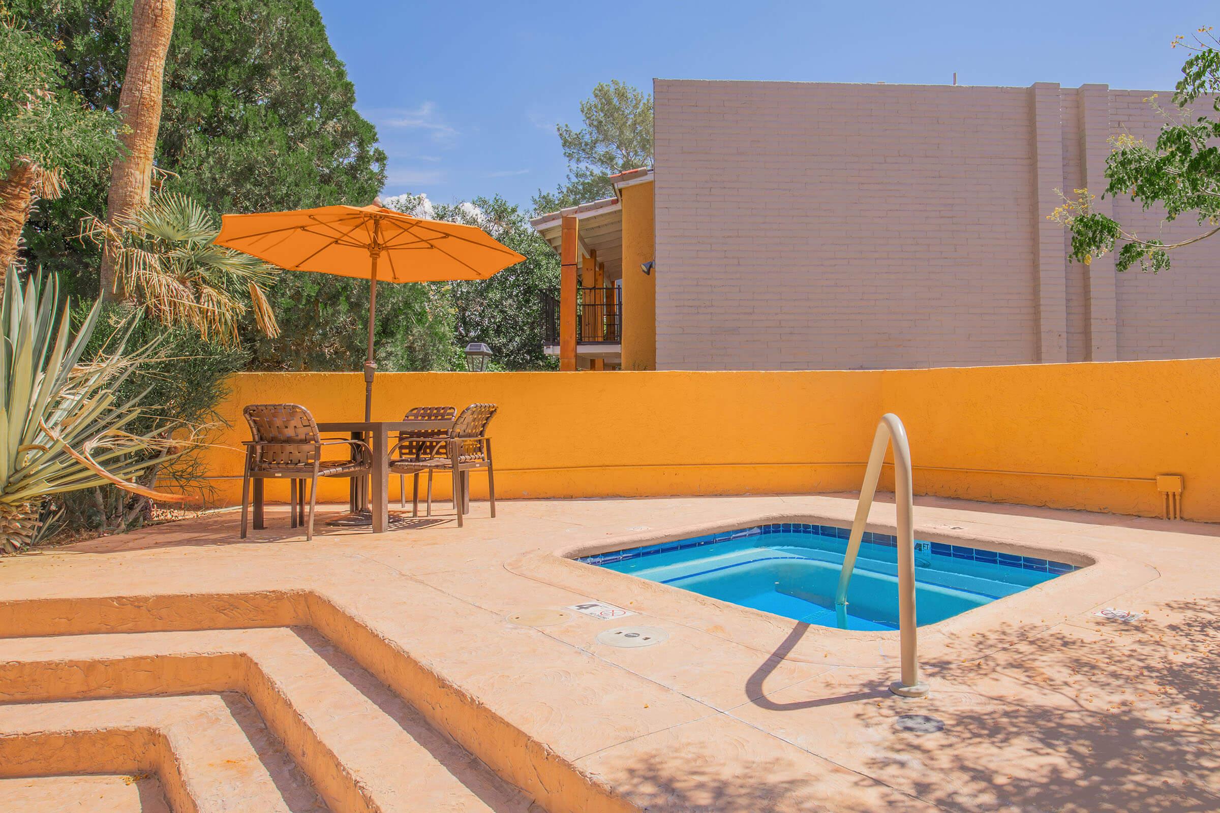 A cozy outdoor space featuring a small hot tub surrounded by a textured patio. There is a round table with four chairs shaded by an orange umbrella. Lush greenery and palm trees are present, adding to the serene atmosphere, with a beige building visible in the background against a clear blue sky.