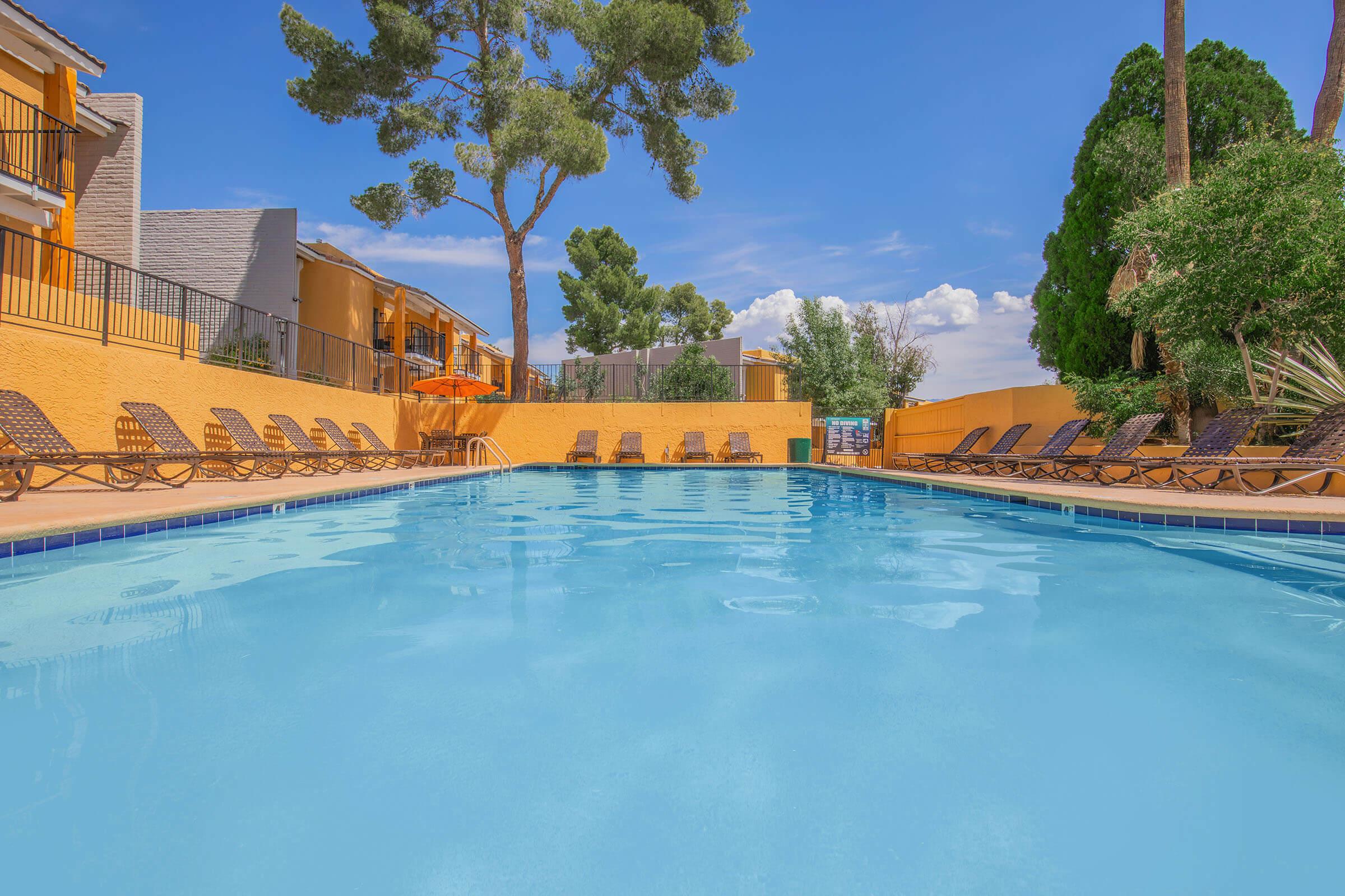 A swimming pool surrounded by lounge chairs, set against a bright blue sky with a few clouds. Green trees are visible in the background, and colorful buildings flank the pool area, suggesting a relaxing outdoor space.