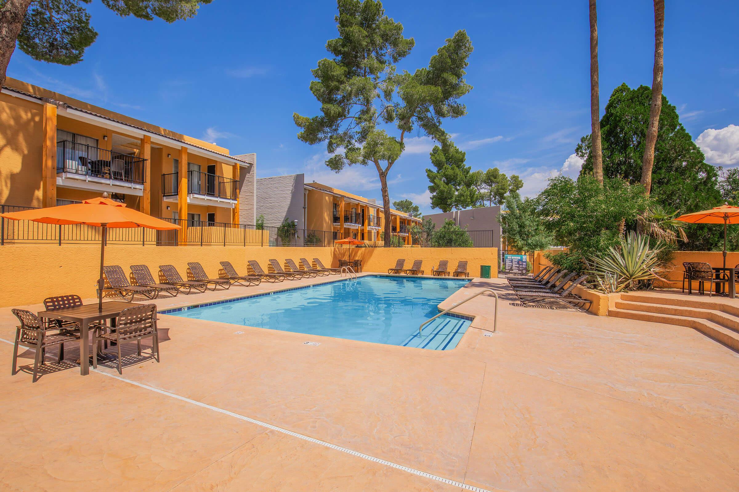 A sunny outdoor pool area surrounded by lounge chairs, with a warm orange-colored deck. Spacious greenery and tall trees provide shade nearby. Bright orange umbrellas offer additional shade, while multicolored outer buildings are in the background under a clear blue sky.
