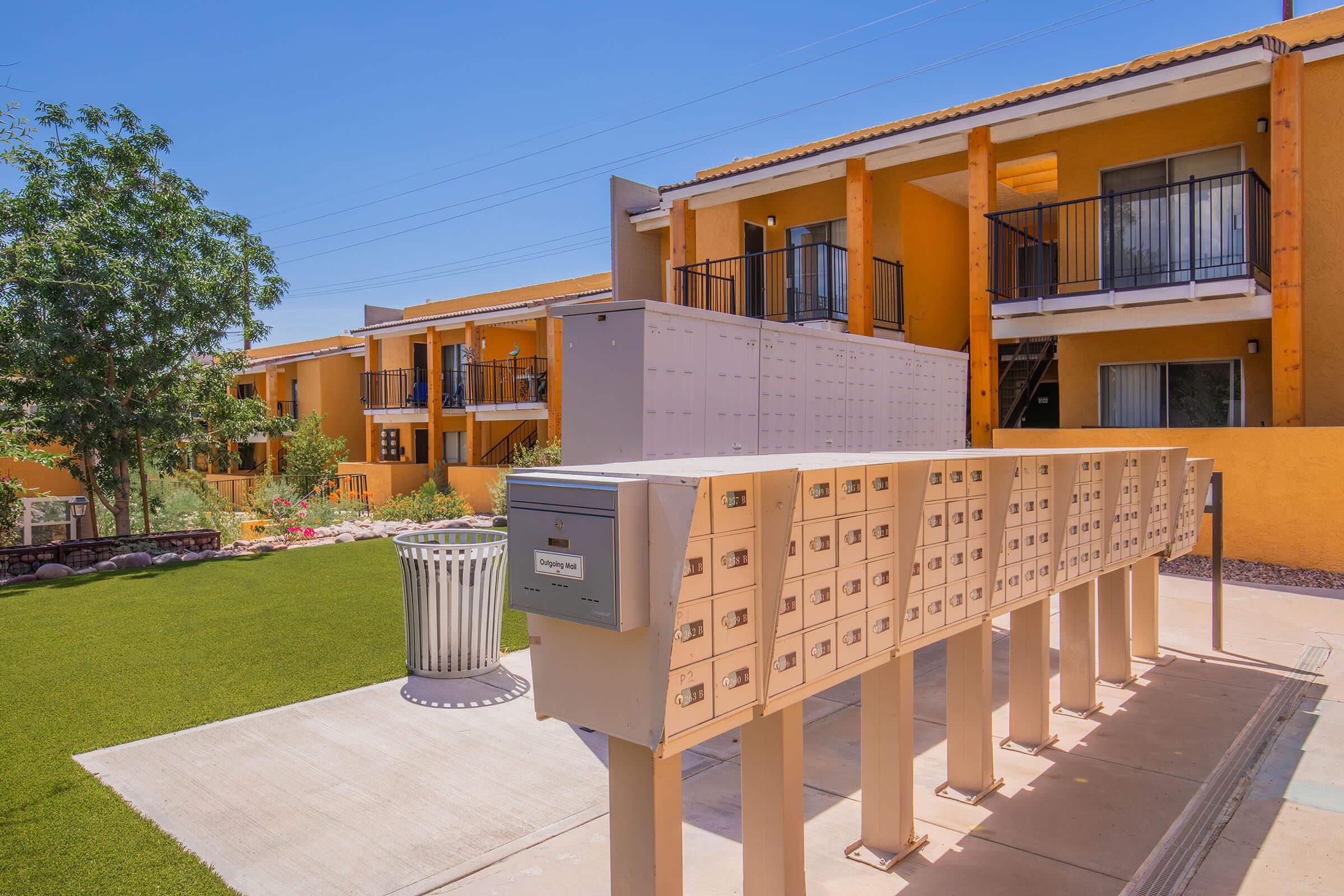 A row of mailbox units in front of a residential building. The setting features well-maintained landscaping with green grass, a tree, and a trash bin. The buildings in the background are painted in warm orange hues, and there are balconies visible on the upper floors. The scene is bright and sunny.