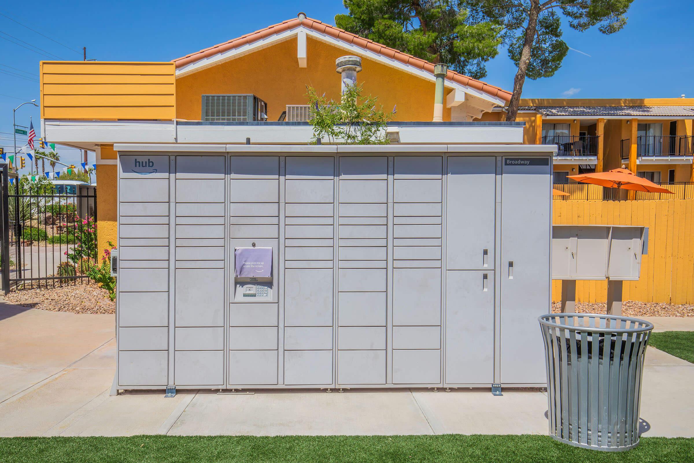 A modern parcel locker station with multiple compartments, situated outdoors near a residential building. The station is gray with a white panel displaying the brand "hub." A nearby trash can sits on a green lawn. In the background, a yellow building and trees are visible under a clear blue sky.