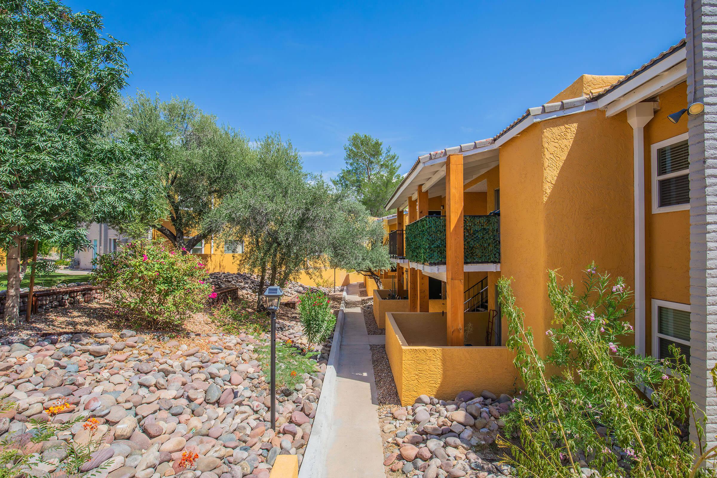 A view of a colorful apartment complex surrounded by well-maintained landscaping, featuring concrete pathways and a variety of plants, including flowering shrubs and stone features. The bright yellow walls of the buildings contrast with the blue sky, creating an inviting outdoor atmosphere.