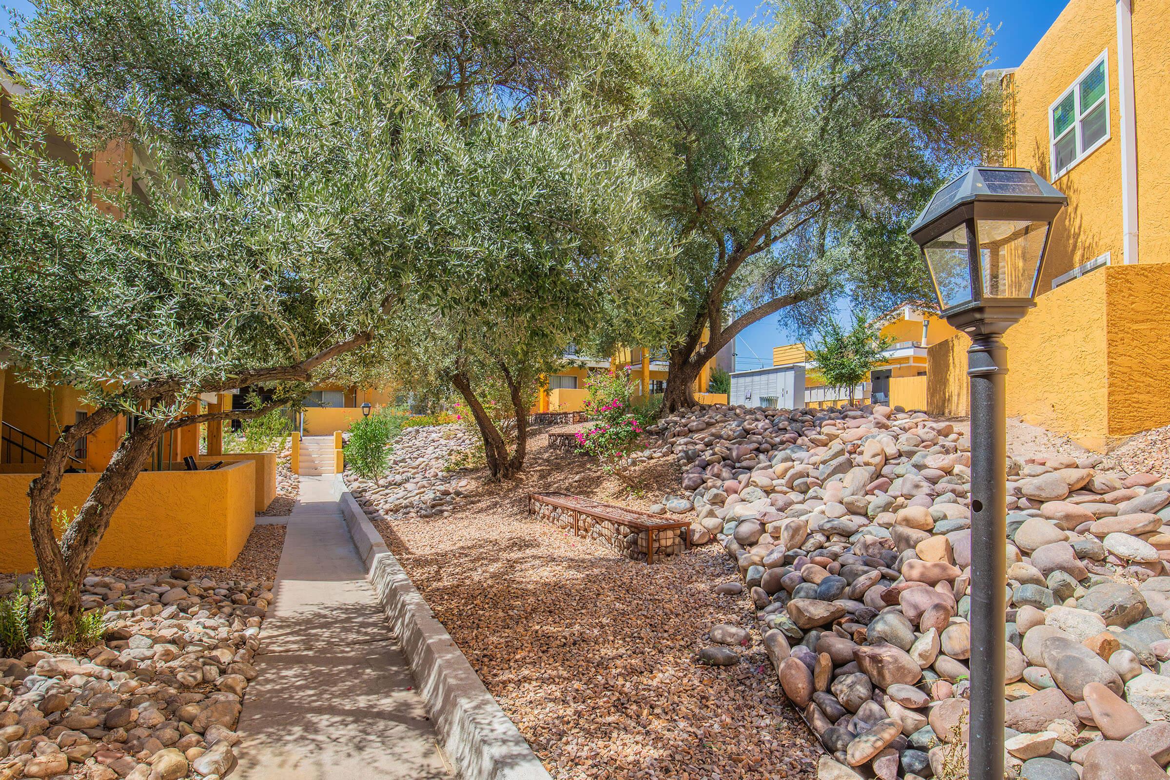 A well-maintained outdoor area featuring a pathway surrounded by large rocks, plants, and trees. A decorative lamp post is visible along the path, which leads through a vibrant landscape of yellow buildings and greenery under a clear blue sky.