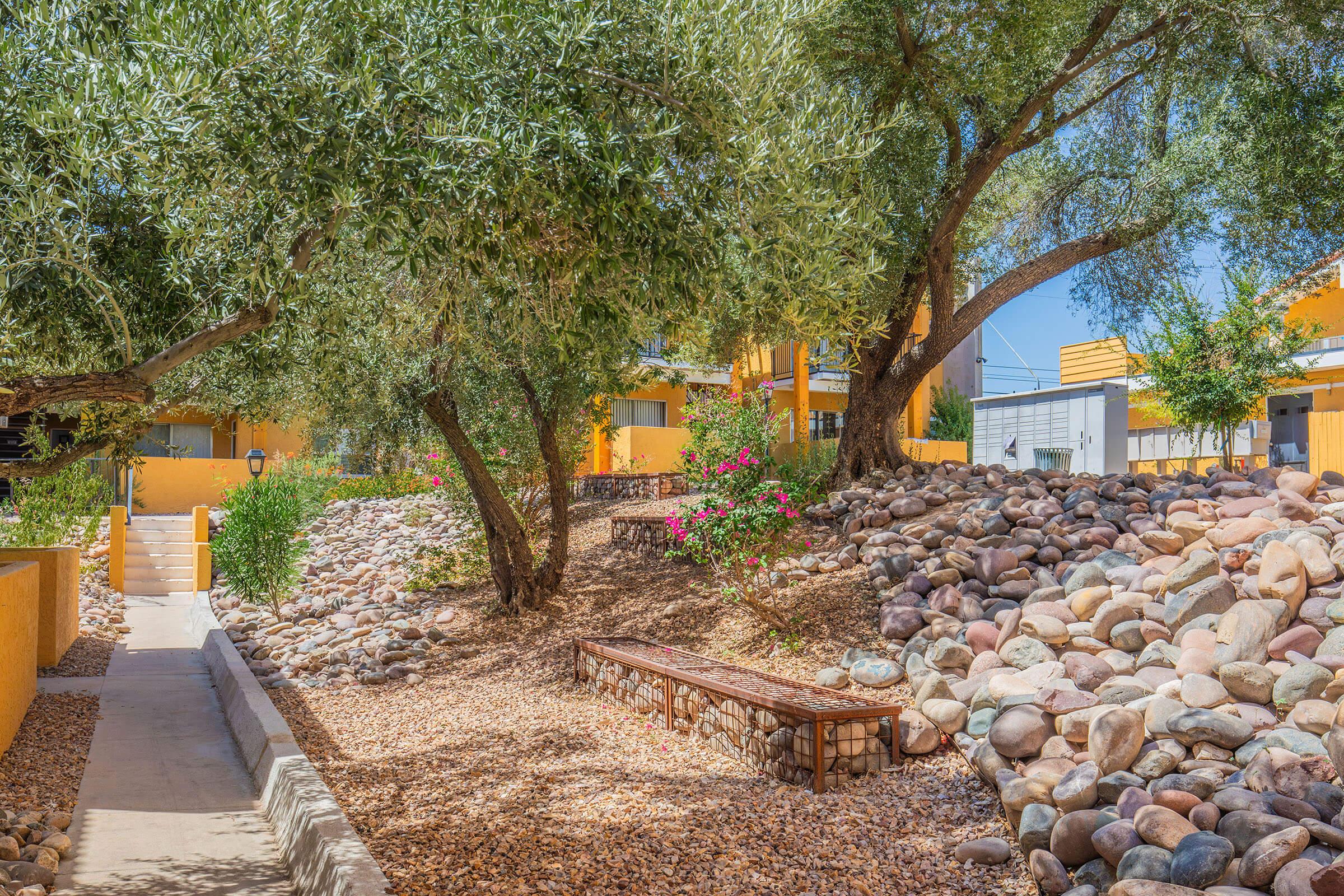 A landscaped path featuring smooth stones, benches, and vibrant greenery. Olive trees and flowering bushes create a serene atmosphere, while brightly colored buildings are partially visible in the background under a clear blue sky.