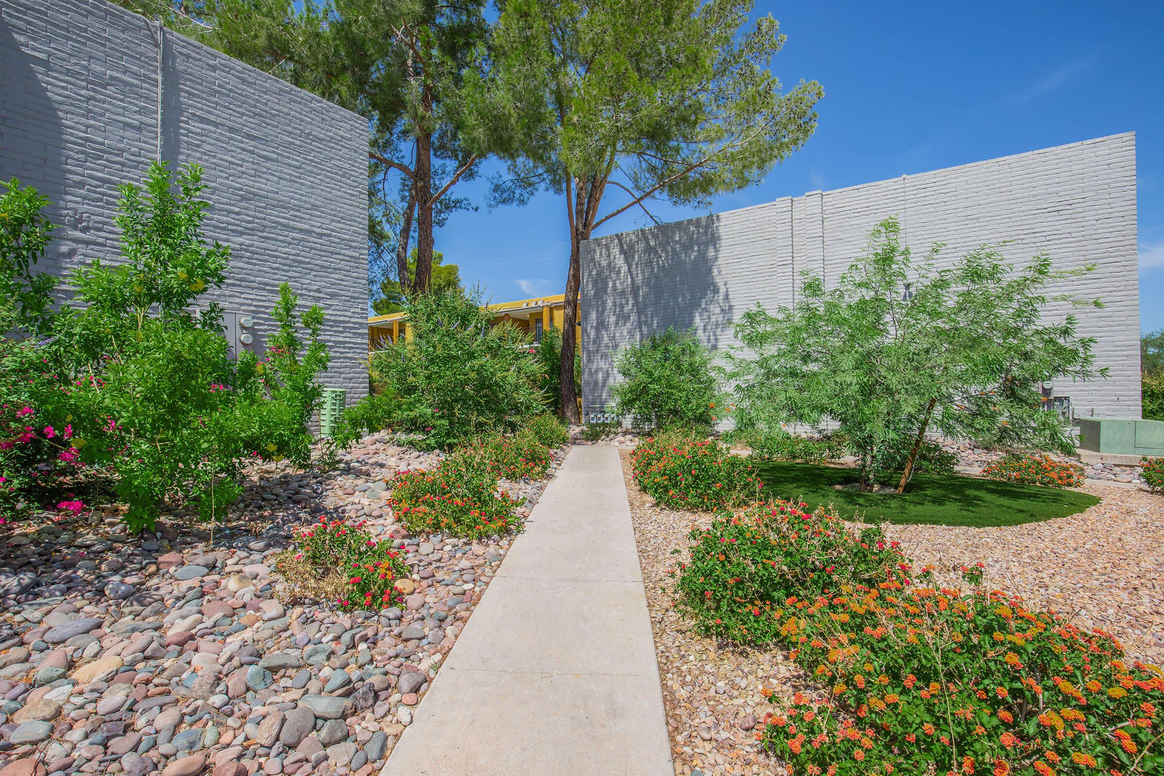 A landscaped garden featuring a stone pathway lined with vibrant flowers and greenery. Tall trees provide shade, with textured grey walls framing the scene under a clear blue sky. The design offers a peaceful outdoor environment.