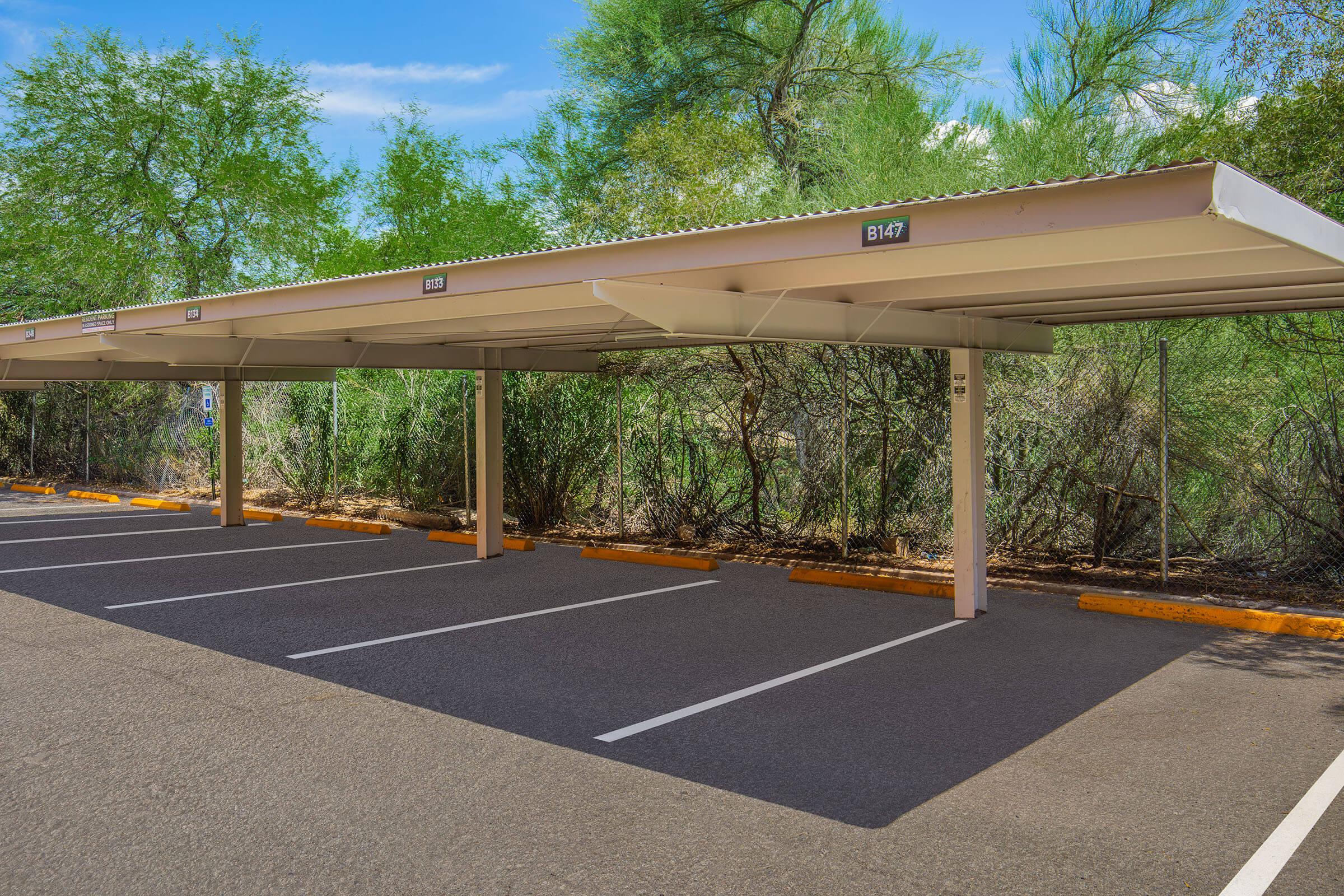 Covered parking spaces with numbered signs in a well-maintained lot. Lush green vegetation and trees are in the background. The asphalt surface is clean, and the area is well-lit by natural sunlight.