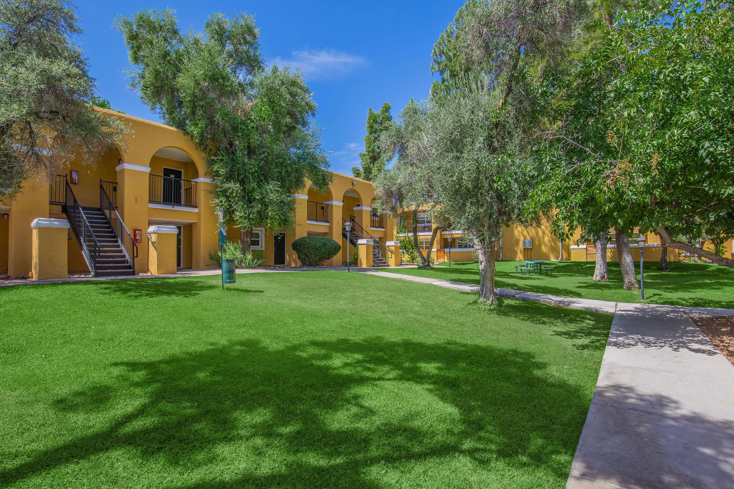 A sunny courtyard featuring well-maintained grass and trees, with yellow apartment buildings in the background. There are two sets of stairs leading to the entrances of the apartments, and a pathway runs through the green space, creating a pleasant and inviting atmosphere.