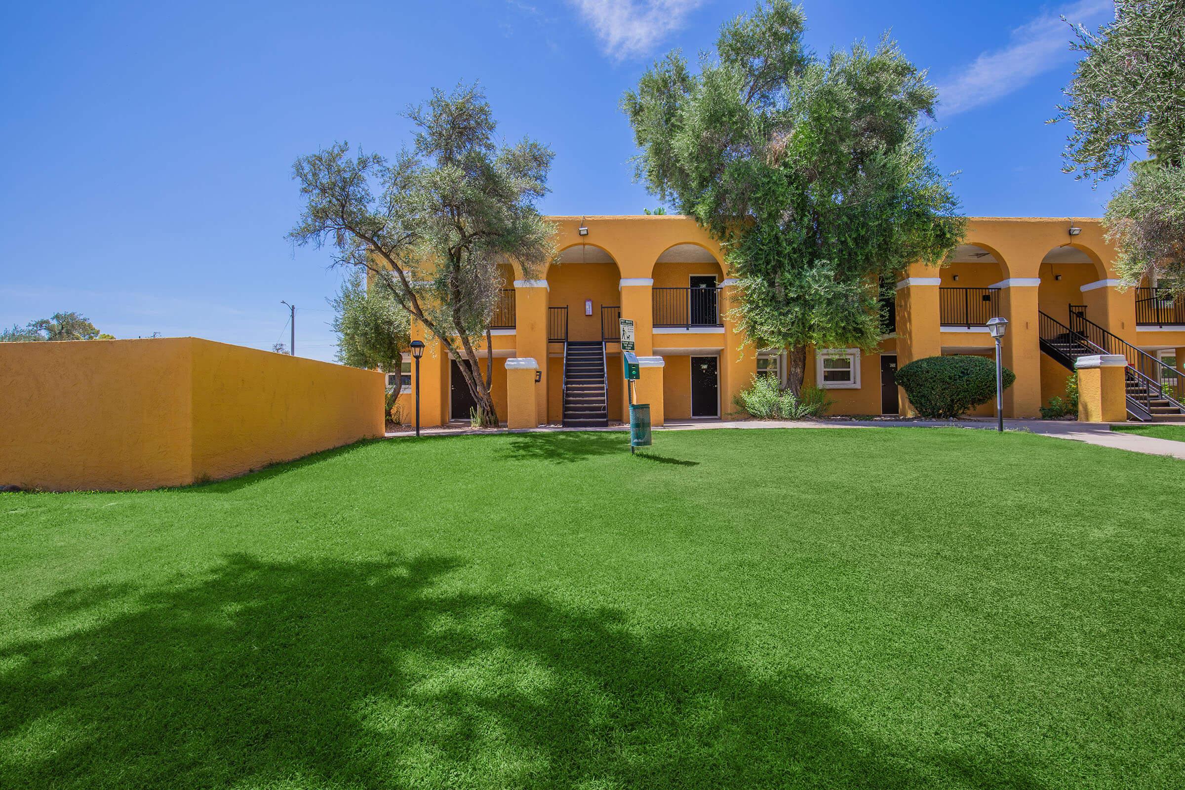 A bright yellow apartment building with arched entrances, surrounded by lush green grass and a few trees. There is a signpost on the lawn, and the sky is clear with some clouds.