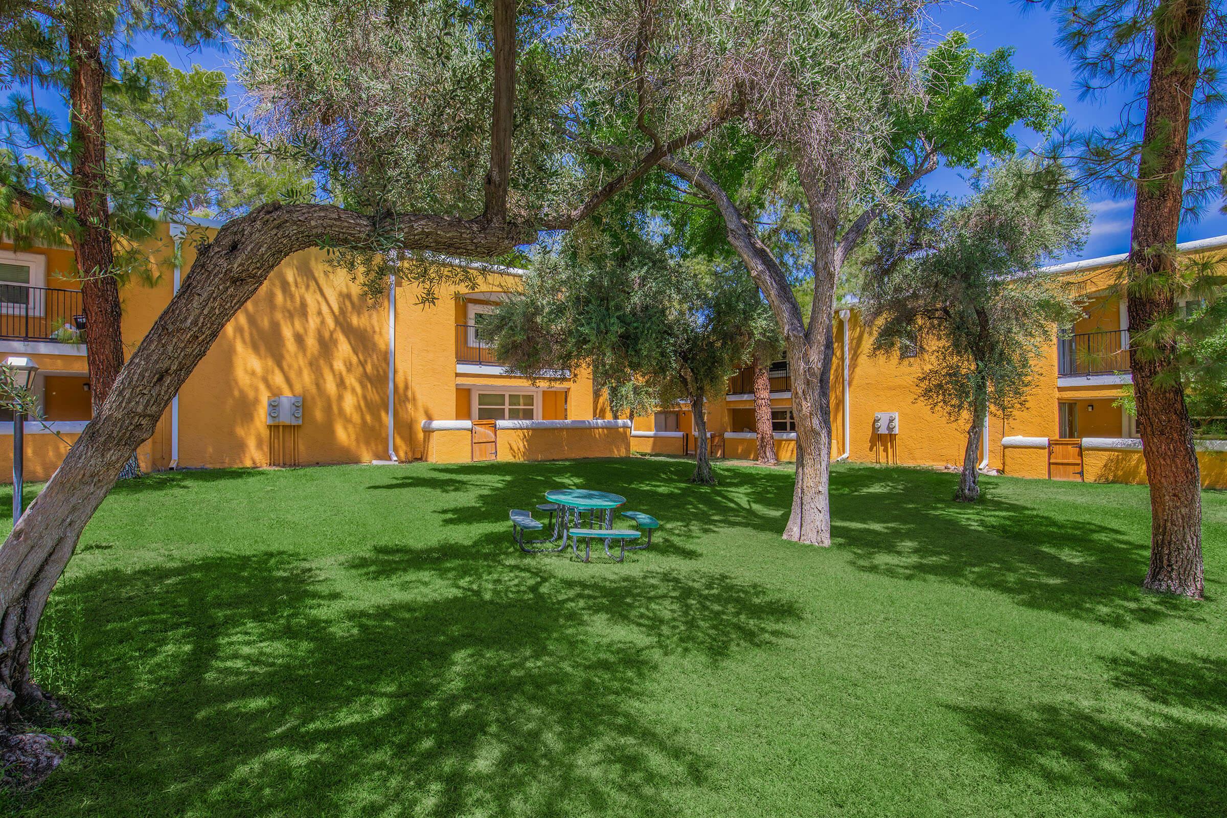 A grassy area with a round blue table and chairs surrounded by trees, set in front of a building with yellow walls. The scene is bright and sunny, showcasing a peaceful outdoor space ideal for relaxation.