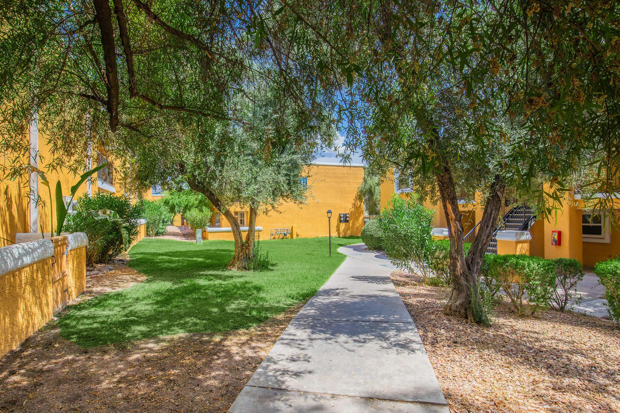 Lush green pathway winding through a landscaped area with trees and shrubs, leading to orange-colored buildings. Bright sunlight filters through the foliage, creating a serene and inviting atmosphere. The scene suggests a peaceful outdoor space, ideal for relaxation or strolling.