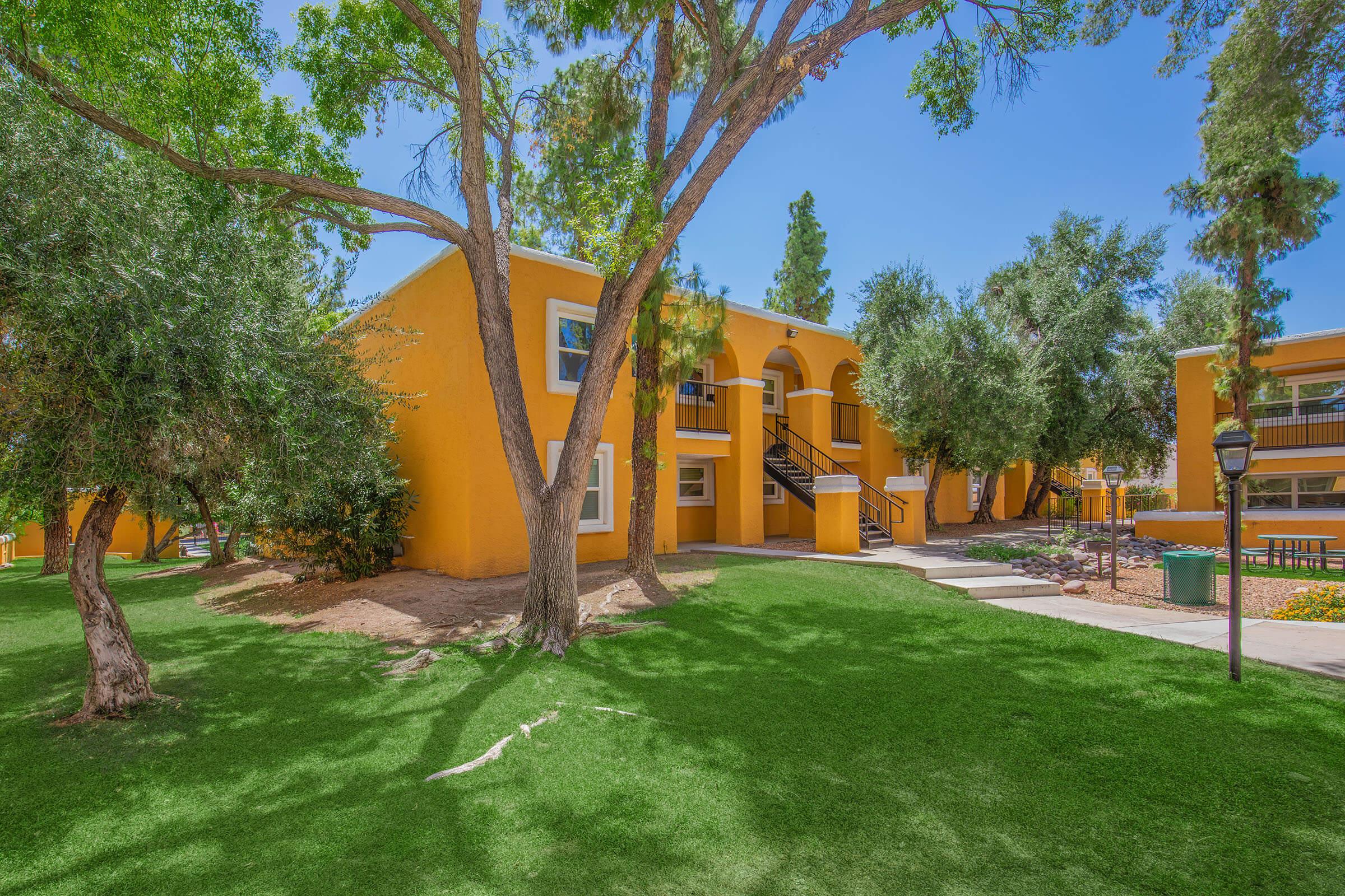 A colorful yellow building surrounded by green grass and trees. The building has an arched entrance and multiple windows. There are pathways leading through the lush landscape, and outdoor seating areas are visible nearby, creating a vibrant and welcoming atmosphere.