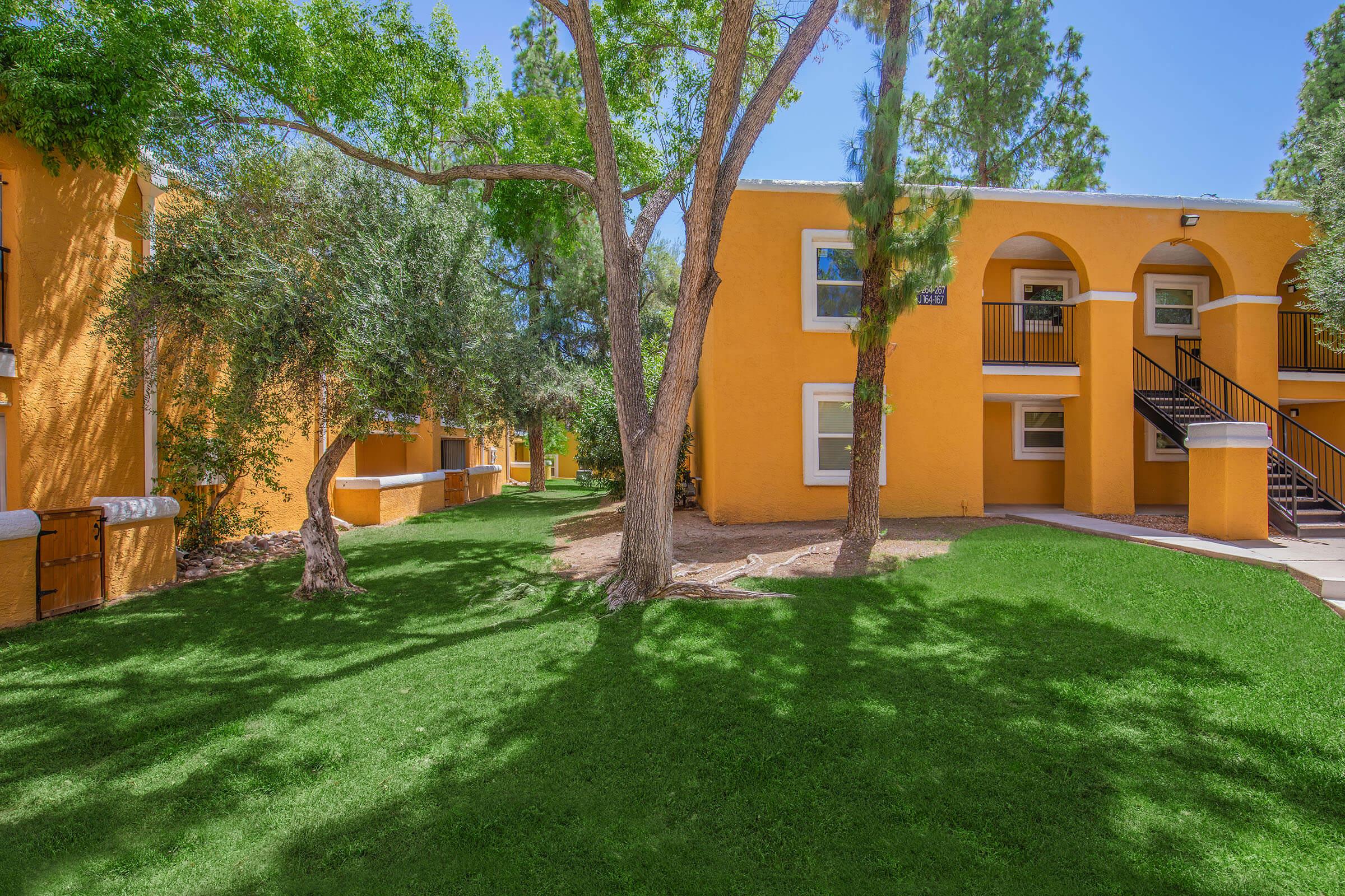 A bright, sunny outdoor scene featuring a well-maintained courtyard with lush green grass, trees, and vibrant yellow-orange apartment buildings. The setting appears inviting and tranquil, perfect for relaxation or outdoor activities.