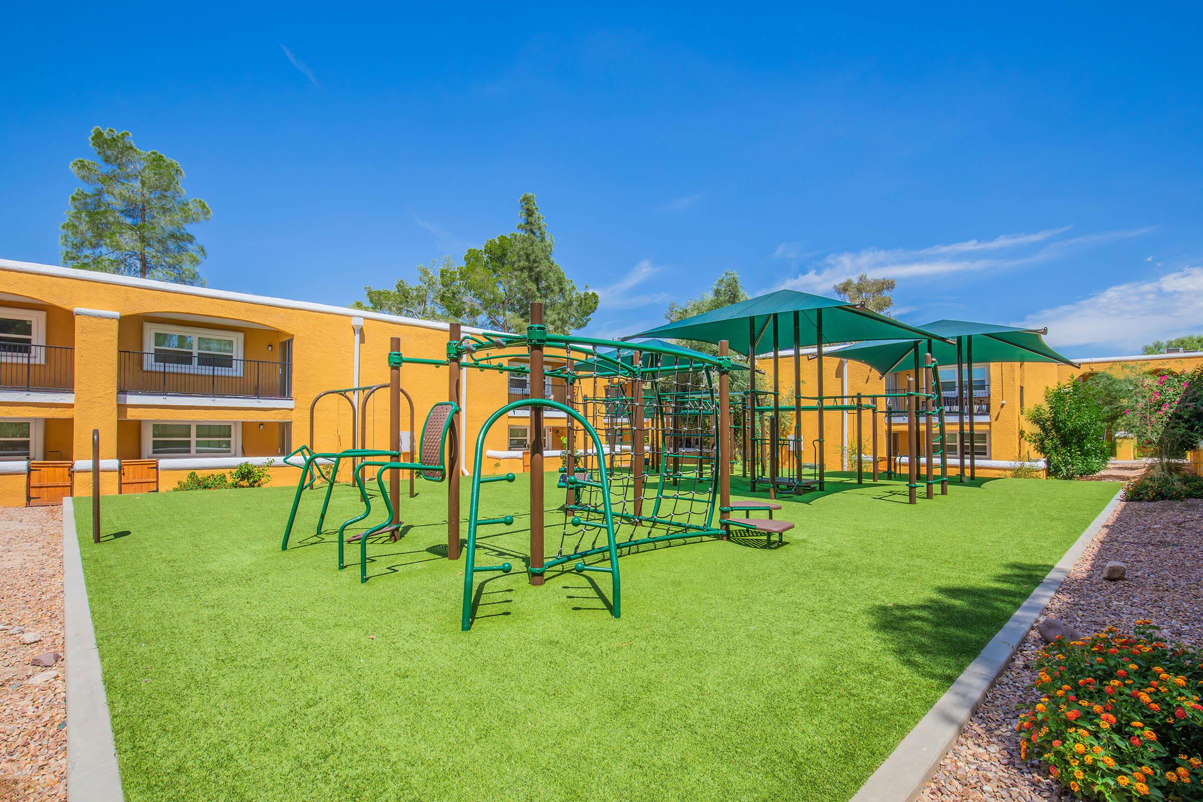 A vibrant outdoor playground set on artificial grass, featuring climbing structures and swings. The playground is surrounded by colorful landscaping and bordered by two yellow apartment buildings under a clear blue sky, creating a cheerful and inviting atmosphere for children.