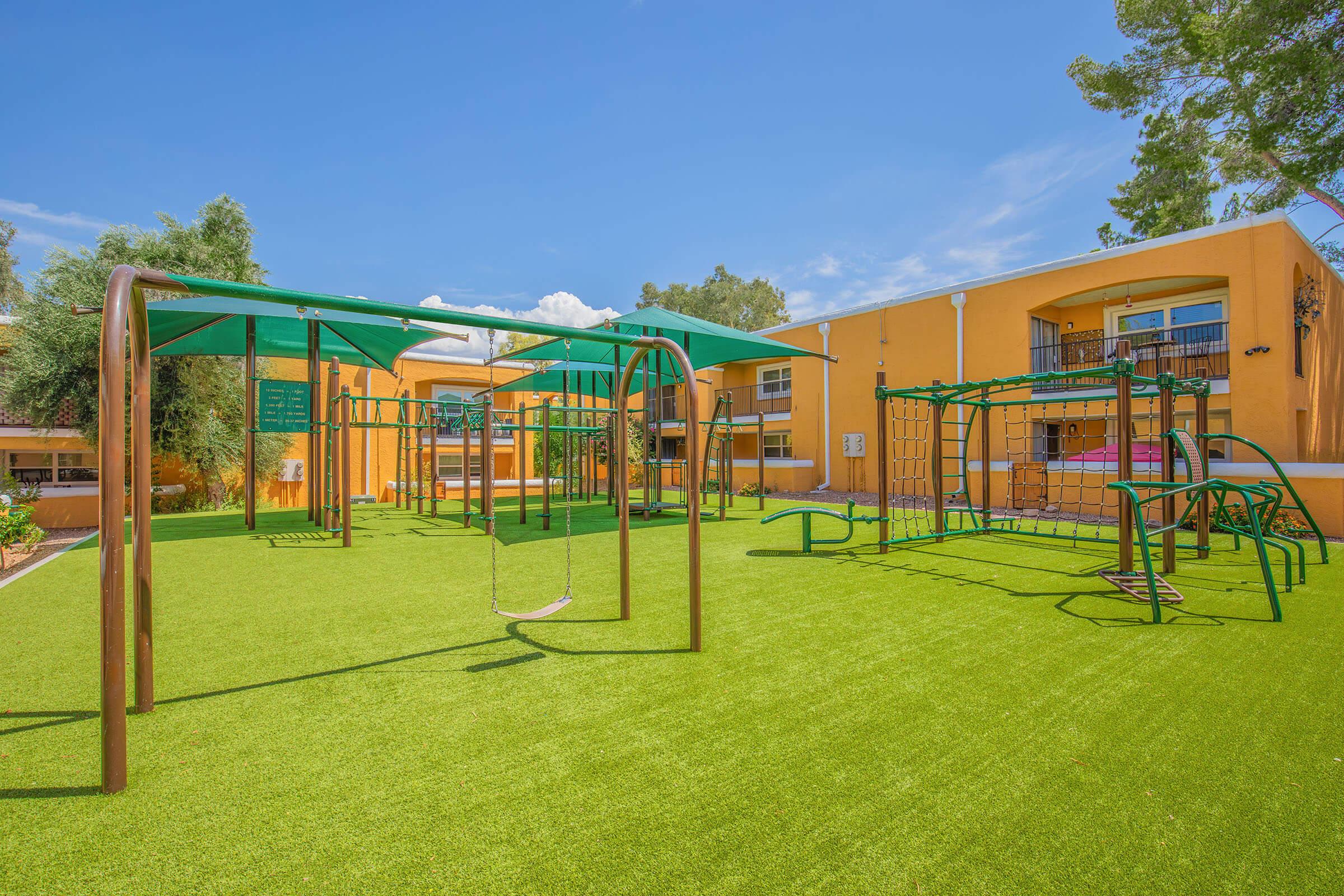 Playground with green synthetic grass featuring swings, climbing structures, and monkey bars, surrounded by colorful buildings and trees under a clear blue sky. Ideal for children's outdoor play and physical activity.