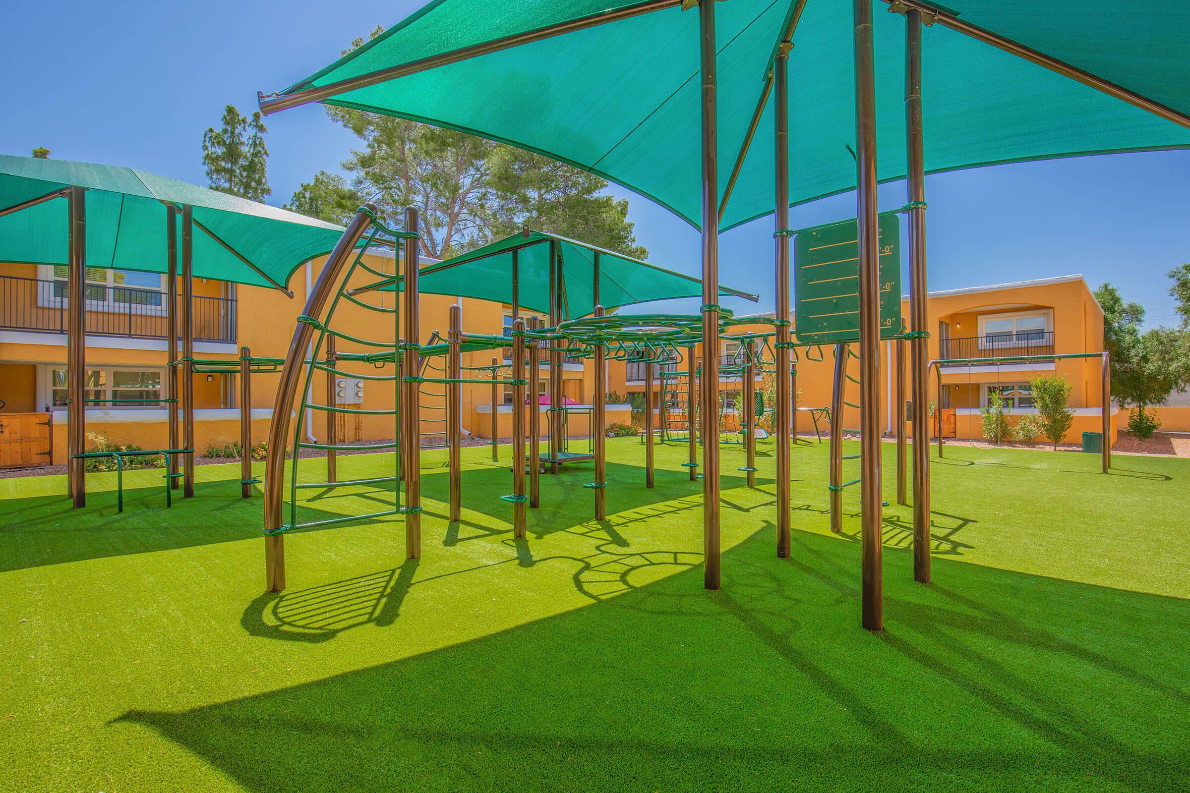 Playground with green artificial turf and various climbing structures under shade canopies. Surrounding buildings are painted in warm colors, providing a cheerful and inviting atmosphere. Lush trees in the background enhance the outdoor setting, making it a vibrant area for children to play.