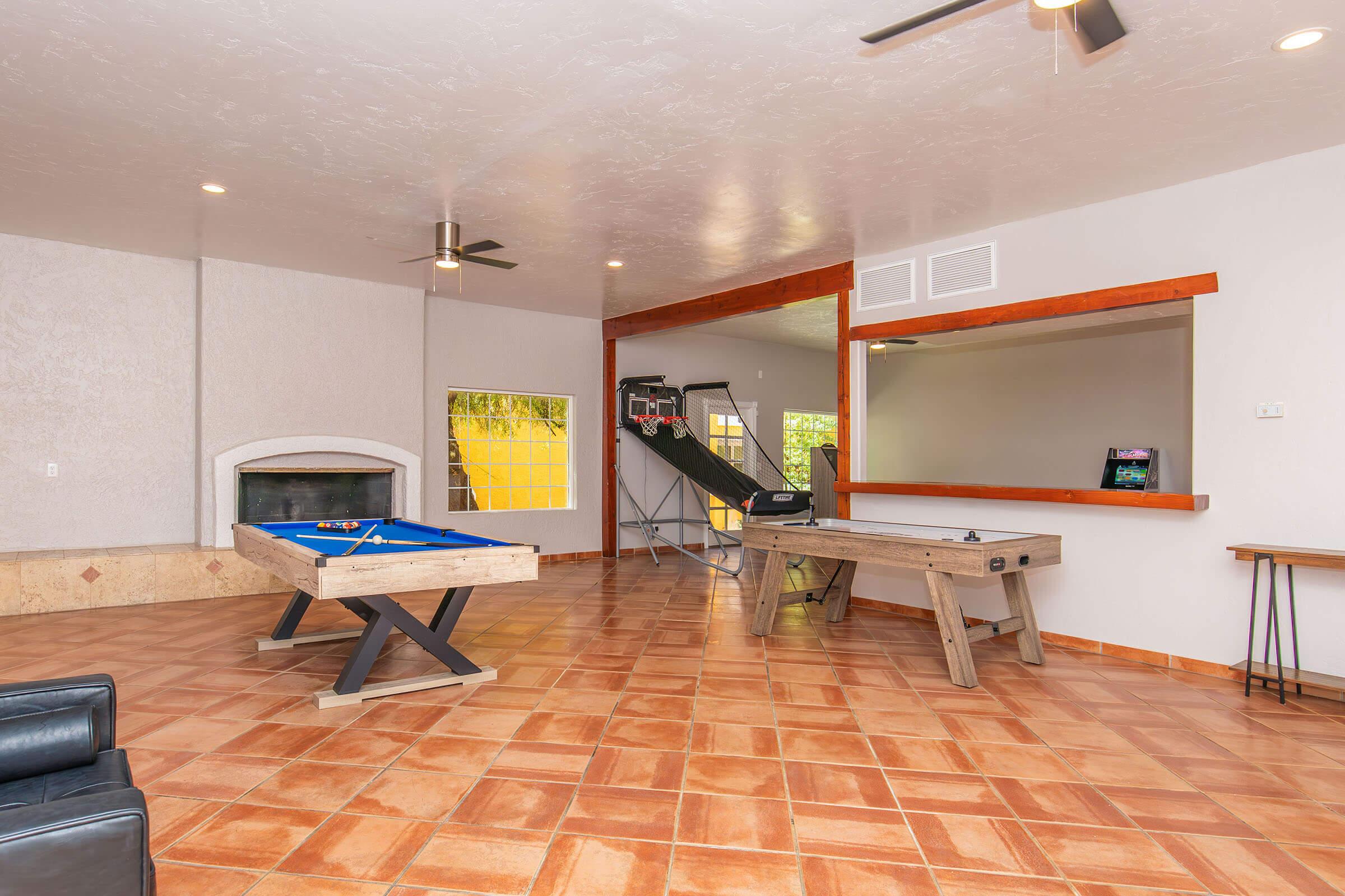 A spacious game room featuring a blue pool table and a wooden table, with a fireplace in the background. Two ceiling fans are visible, along with a basketball hoop and slide area. The room has tiled flooring and large windows allowing natural light to enter.