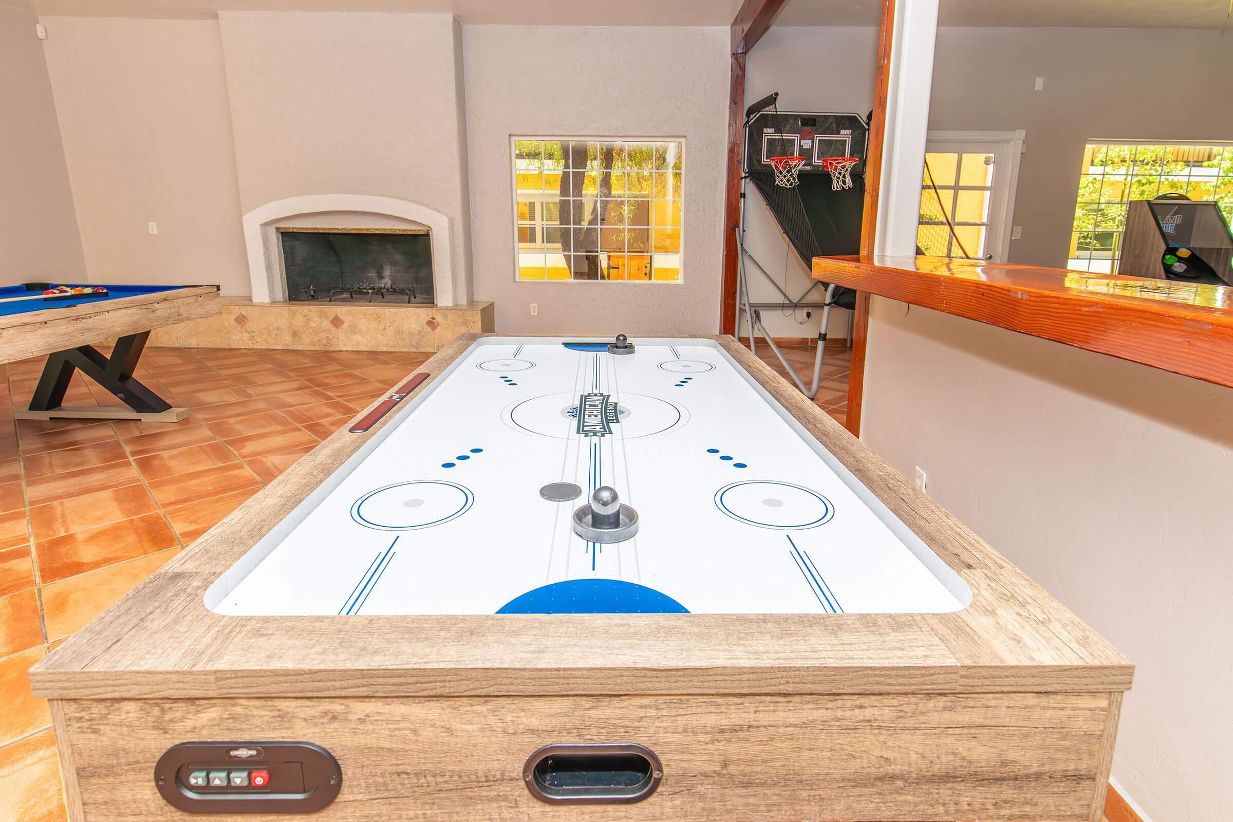A close-up view of an air hockey table with a wooden frame, positioned in a bright game room that features a fireplace and a basketball hoop in the background. The room has large windows allowing natural light to enter, enhancing the inviting atmosphere.