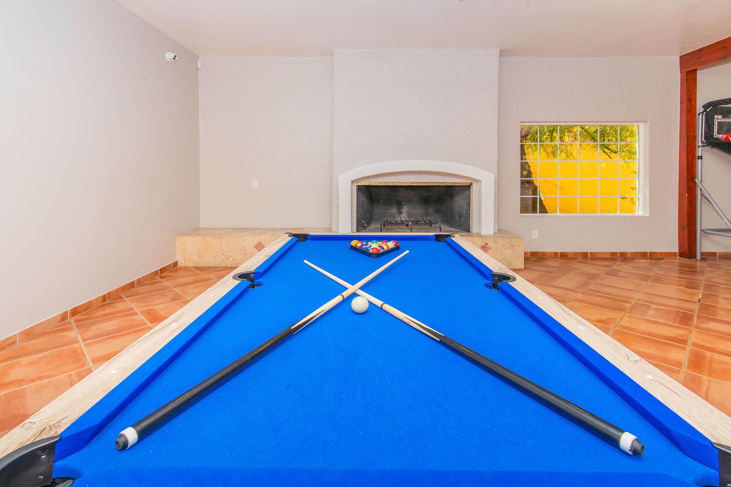 A bright room featuring a blue pool table with two cue sticks crossed over a cue ball. In the background, there is a fireplace and a large window allowing natural light to enter, creating a cozy atmosphere. The floor is tiled with warm-colored ceramic tiles.