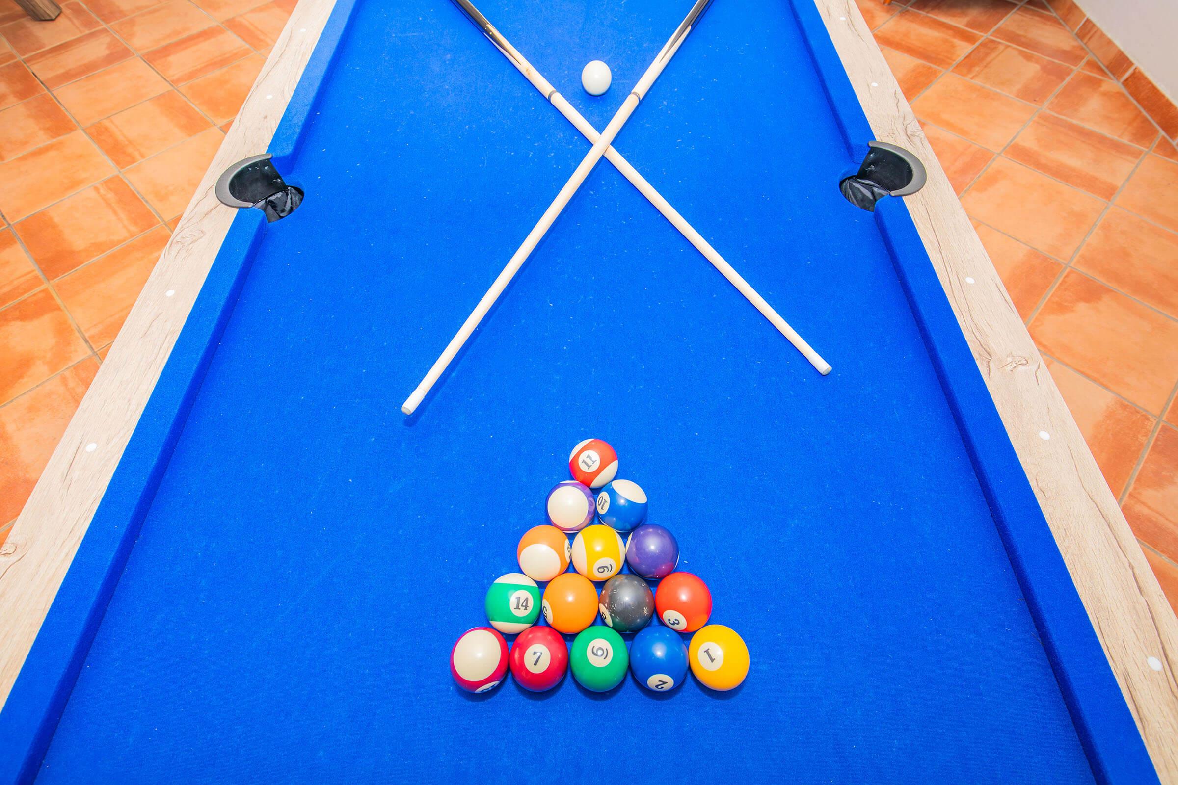 A blue billiard table with a neatly arranged triangle of colored pool balls at the bottom. Two cues are crossed above the balls, with a single white cue ball positioned above them. The floor is tiled in orange hues, creating a warm, inviting atmosphere.