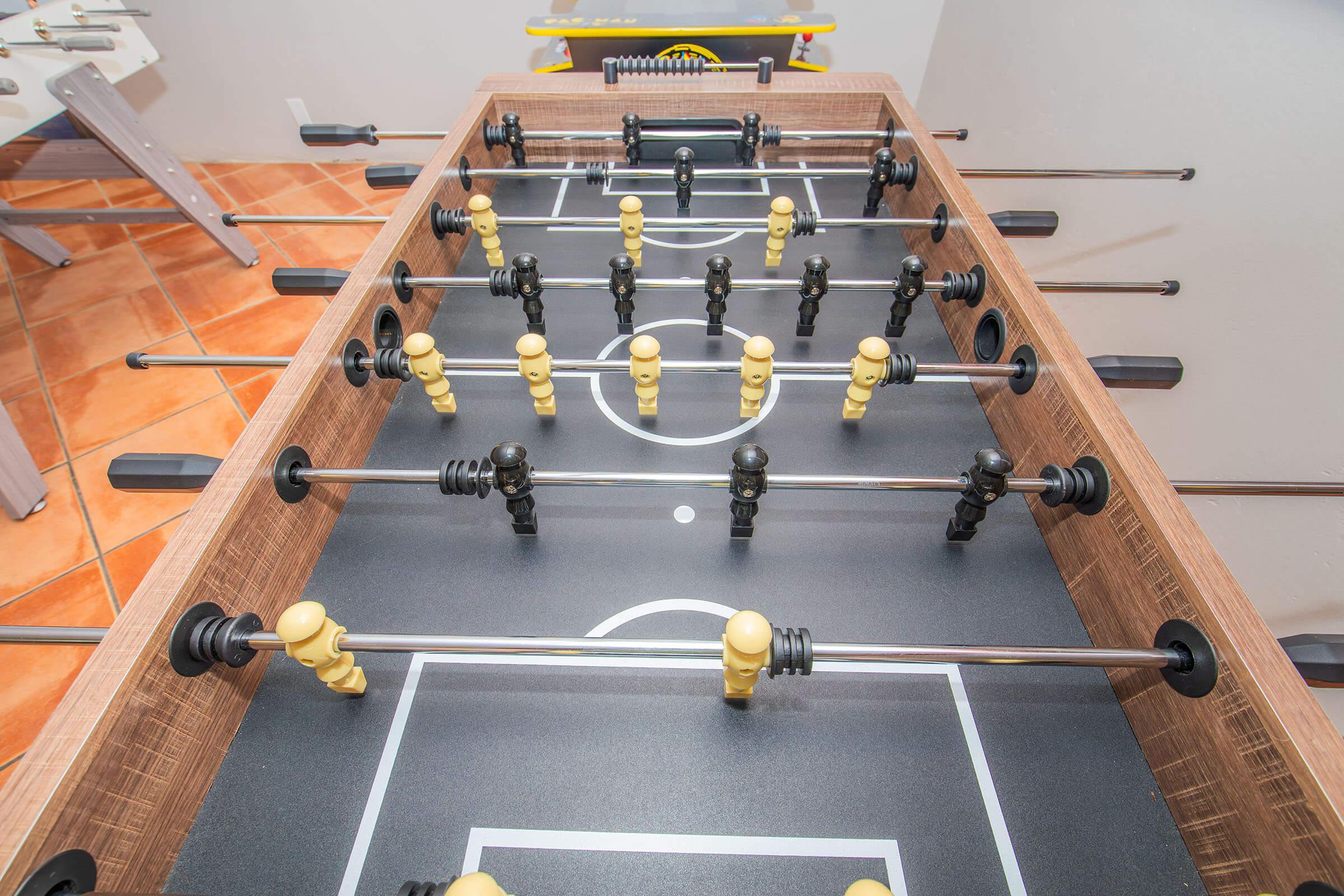 A foosball table with black and yellow player figures arranged on a green field. The table features rods for controlling the players, with a simple layout resembling a soccer pitch, and is set in a well-lit indoor space with tiled flooring.