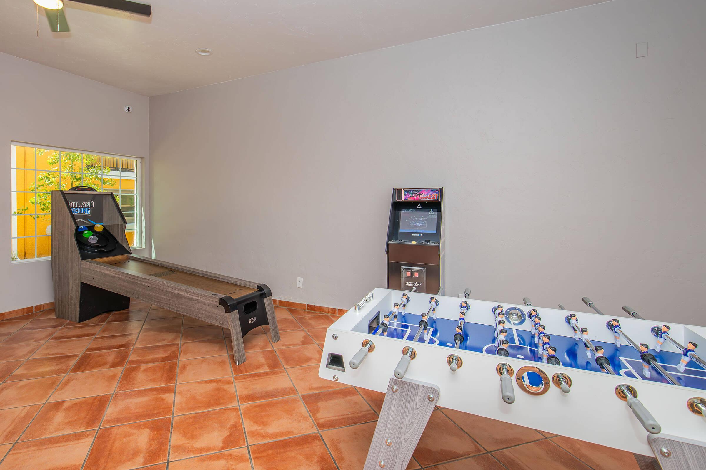 An indoor recreation room featuring a foosball table, a shuffleboard table, and a video game arcade machine. The walls are painted light gray, and the floor is tiled with orange and brown tiles. Large windows allow natural light to enter the space.