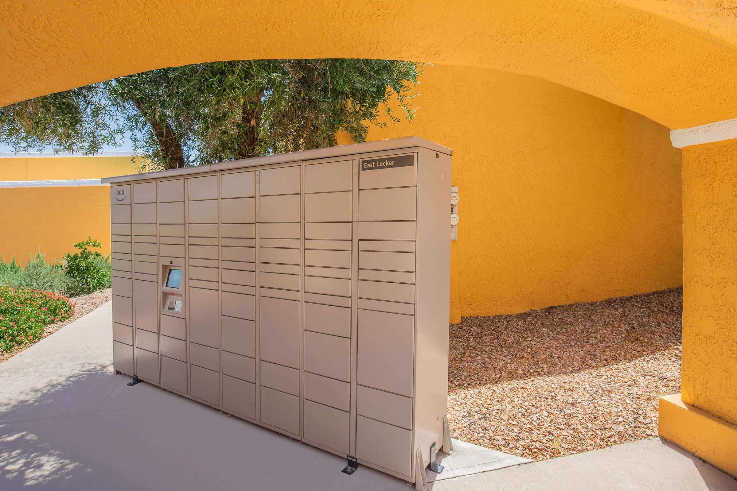 A large, beige package locker stands against a vibrant yellow wall, under an arched structure. Surrounding the locker is a pathway and a gravel area with some greenery, creating a modern and functional outdoor space for package delivery.