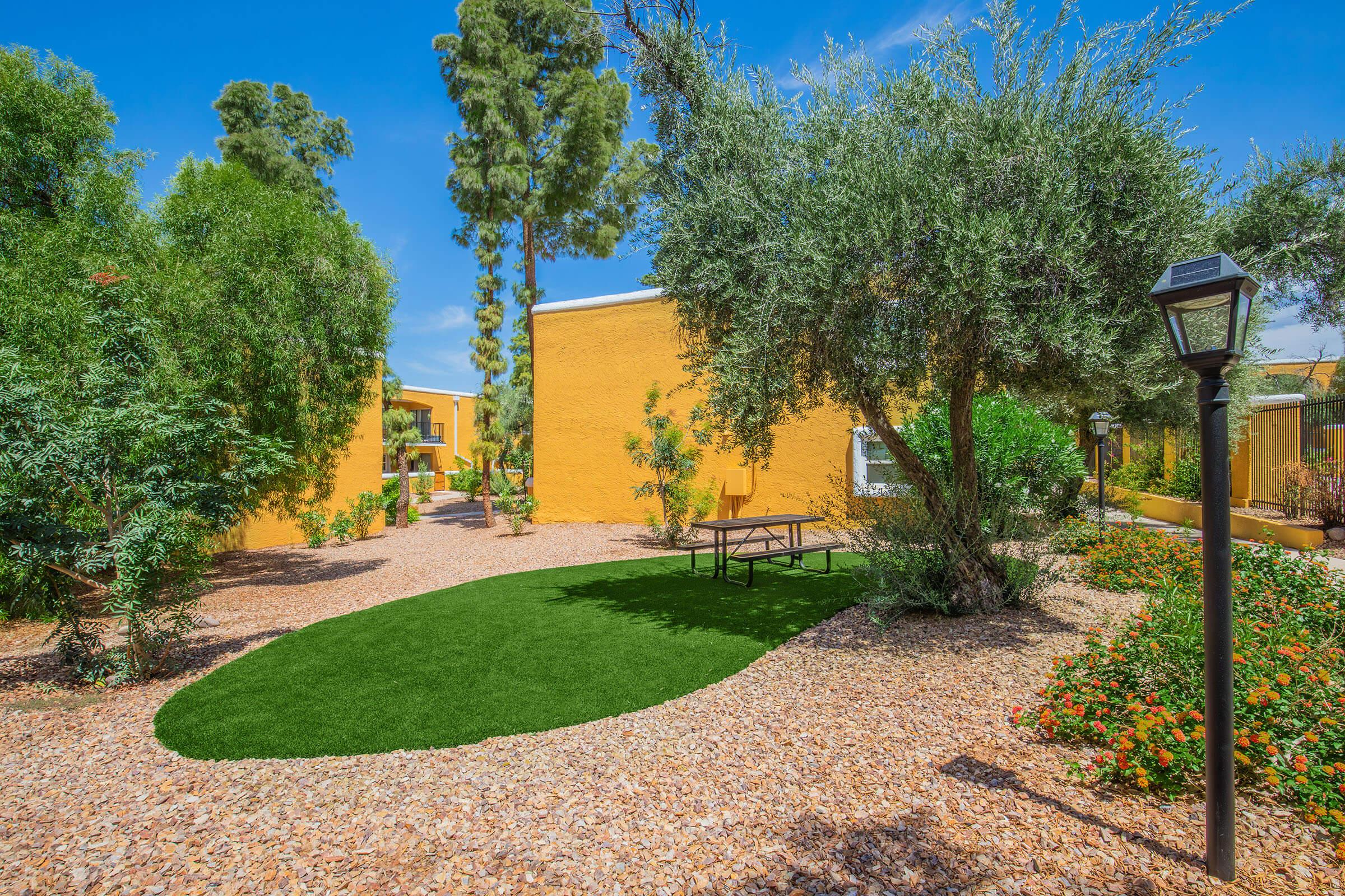 A picturesque outdoor area featuring vibrant green grass, surrounded by small shrubs and flower beds. In the background, there’s a bright yellow building with a pathway leading through the garden. A lamp post stands nearby, adding to the serene atmosphere. Clear blue skies enhance the tranquil setting.