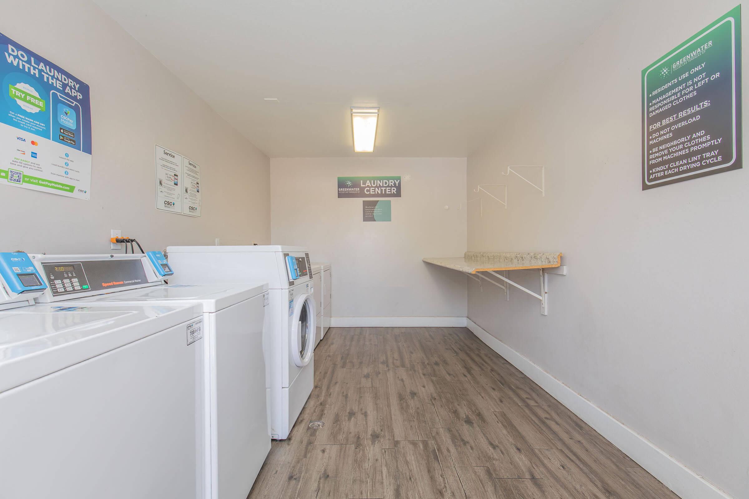 A spacious laundry room featuring multiple washers and dryers, with a folding station along one wall. The walls are painted in light colors, enhancing the bright and organized atmosphere. Informational signs are displayed, providing instructions and details about the laundry facilities.