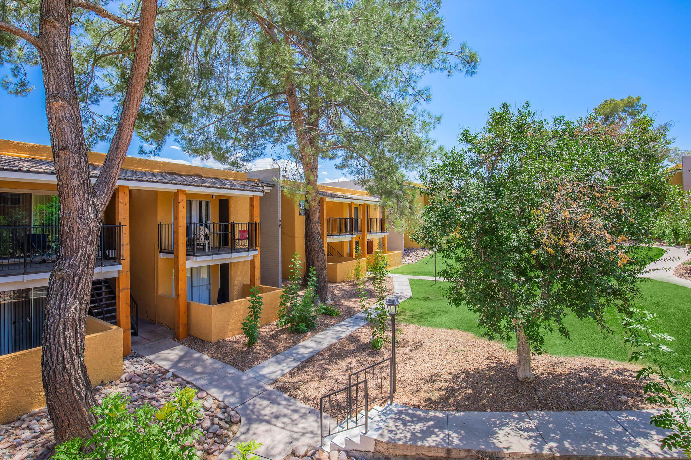 A picturesque view of an apartment complex featuring yellow buildings with balconies, surrounded by lush green trees and landscaped gardens. Pathways wind through the grounds, providing access to the units, under a clear blue sky.