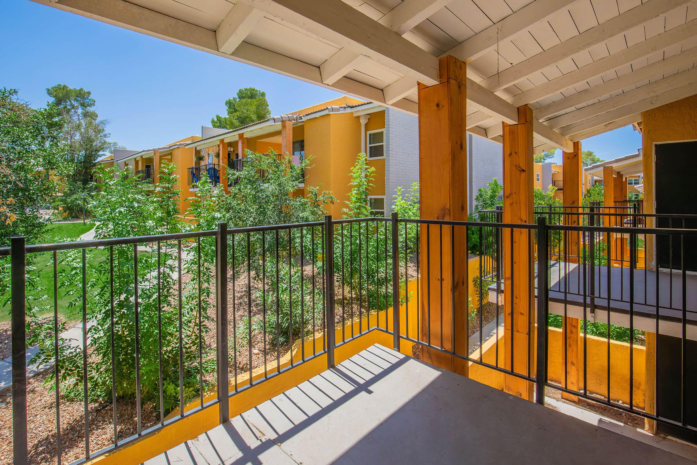 A view from a balcony overlooking a landscaped area with green shrubs and small trees, adjacent to colorful apartment buildings painted in shades of yellow and orange. The railing is black, and the scene is bright and inviting with sunlight casting shadows on the balcony floor.